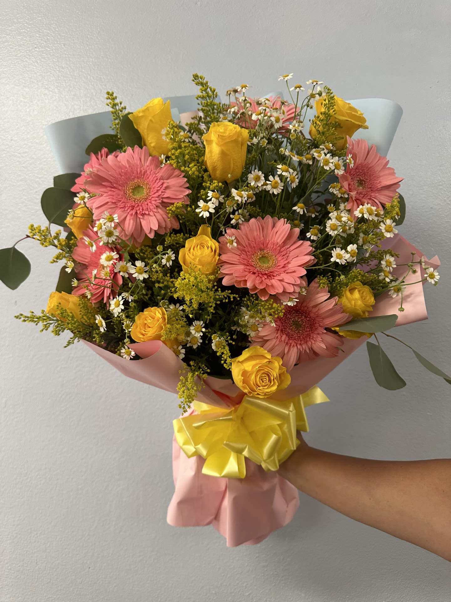 Garden Breeze Bouquet - Bright pink gerbera daisies meet sunny yellow roses in a Garden Breeze bouquet that feels like a spring morning-ideal as a birthday bouquet, a cheerful thank-you gift, or to celebrate any upbeat occasion. This one-sided flower arrangement is designed to present a full, rounded face of blooms to the recipient so she'll feel genuinely seen the moment it's handed to her.  The bouquet pairs large pink gerberas with bright yellow roses and clusters of small white daisy-like filler flowers for lively contrast and soft texture. Fresh green foliage adds depth, while pink and pale blue wrapping paper frame the stems and a yellow ribbon ties everything together for a polished, gift-ready look. Sized to make an immediate impression without overwhelming, it arrives neatly wrapped and ready to present.