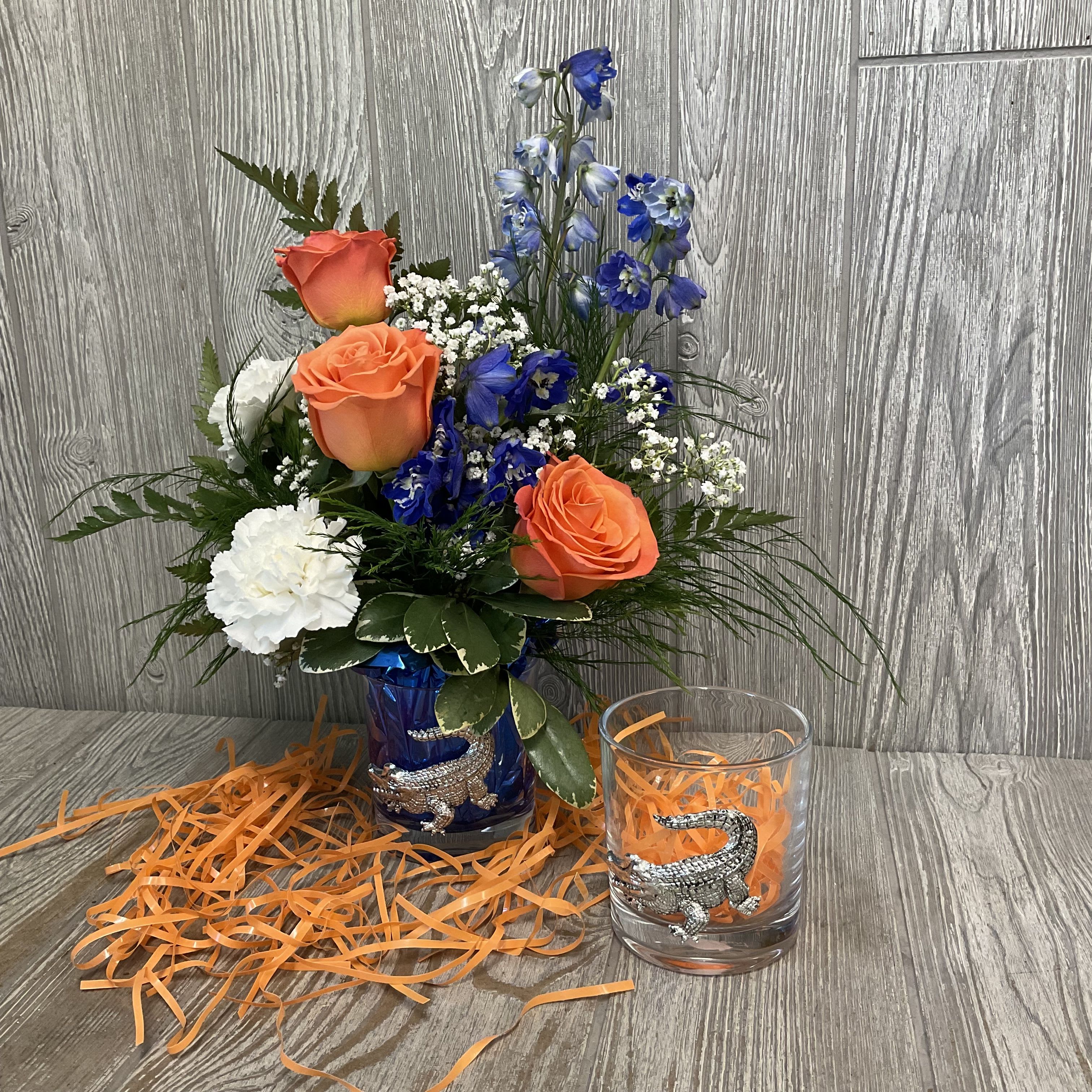 Gator Bar Glass Arraignment  - Three vivid orange roses and tall cobalt-blue delphinium spikes create an immediate, celebratory contrast in this anniversary-ready flower arrangement, perfect for birthdays, graduations, housewarmings, retirements, or simply to say thank you or thinking of you. The playful color story-bright orange, deep blue, crisp white, and fresh green-reads as a joyful, spirited gift that will make the person you're sending it to feel noticed and truly celebrated.  The design pairs three orange roses with white carnations, delicate white filler blooms, and lush greenery, all gathered in a single Arthur Court bar glass vase that amplifies the blue-and-orange palette.