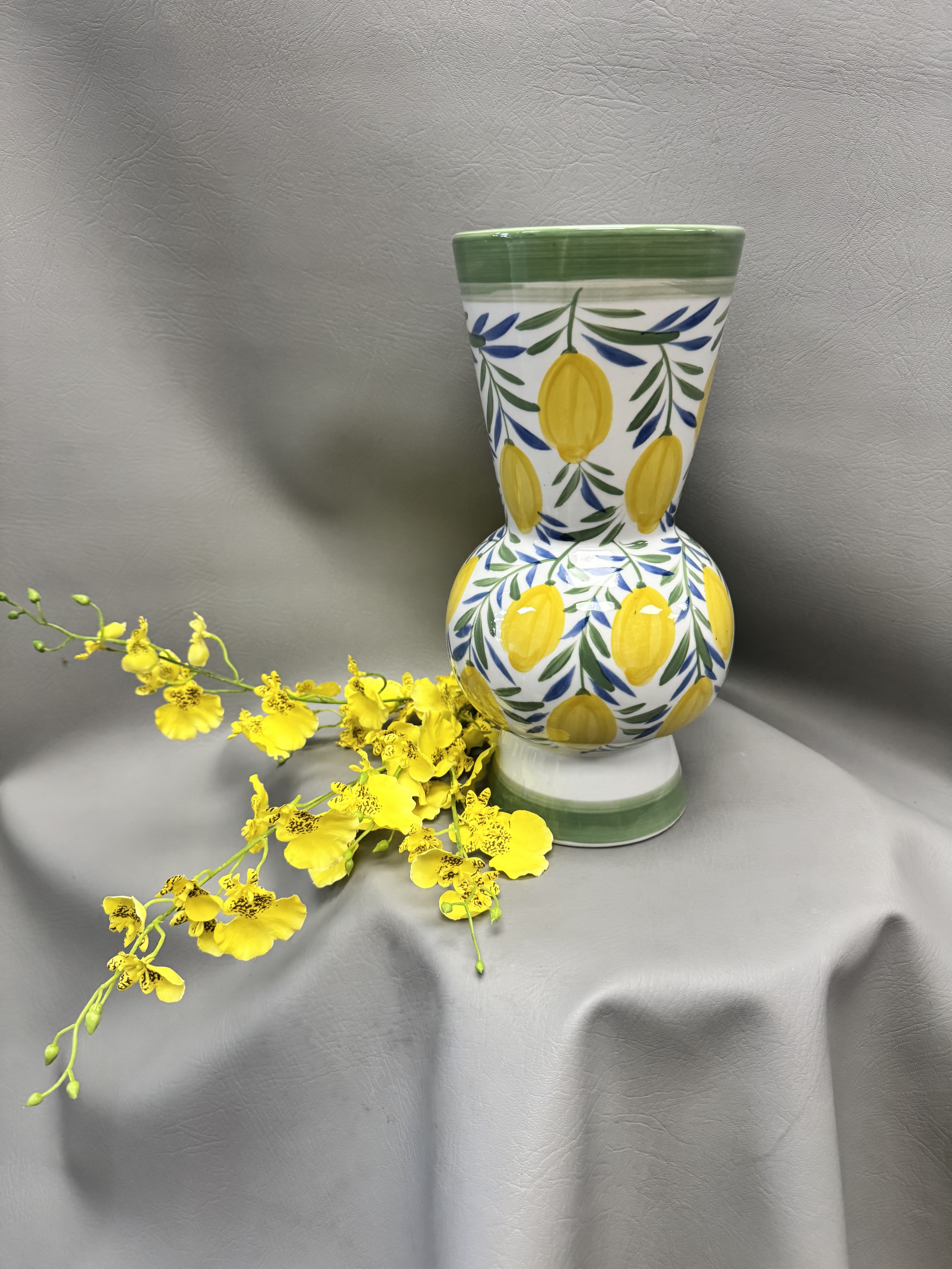 Hand-Painted Lemon Vase.12" H x 6 1/4" Dia - A hand-painted ceramic lemon vase that brings a sunlit, Mediterranean charm to birthdays, housewarmings, or a simple "just because" surprise. The cheerful white body is decorated with yellow fruit-like motifs, blue accents, and a green rim and base that catch the eye and warm a room-perfect for spring decorating or as the centerpiece of a thoughtful thank-you gift.  Measuring 12" H x 6 1/4" diameter, this decorative vase holds fresh or dried stems and also shines as a standalone accent on a mantle, entry table, or kitchen counter. Its trailing, hand-crafted look pairs beautifully with casual or coastal interiors; fill it with seasonal stems, add it to a gift set, or let it stand alone to brighten someone's day.