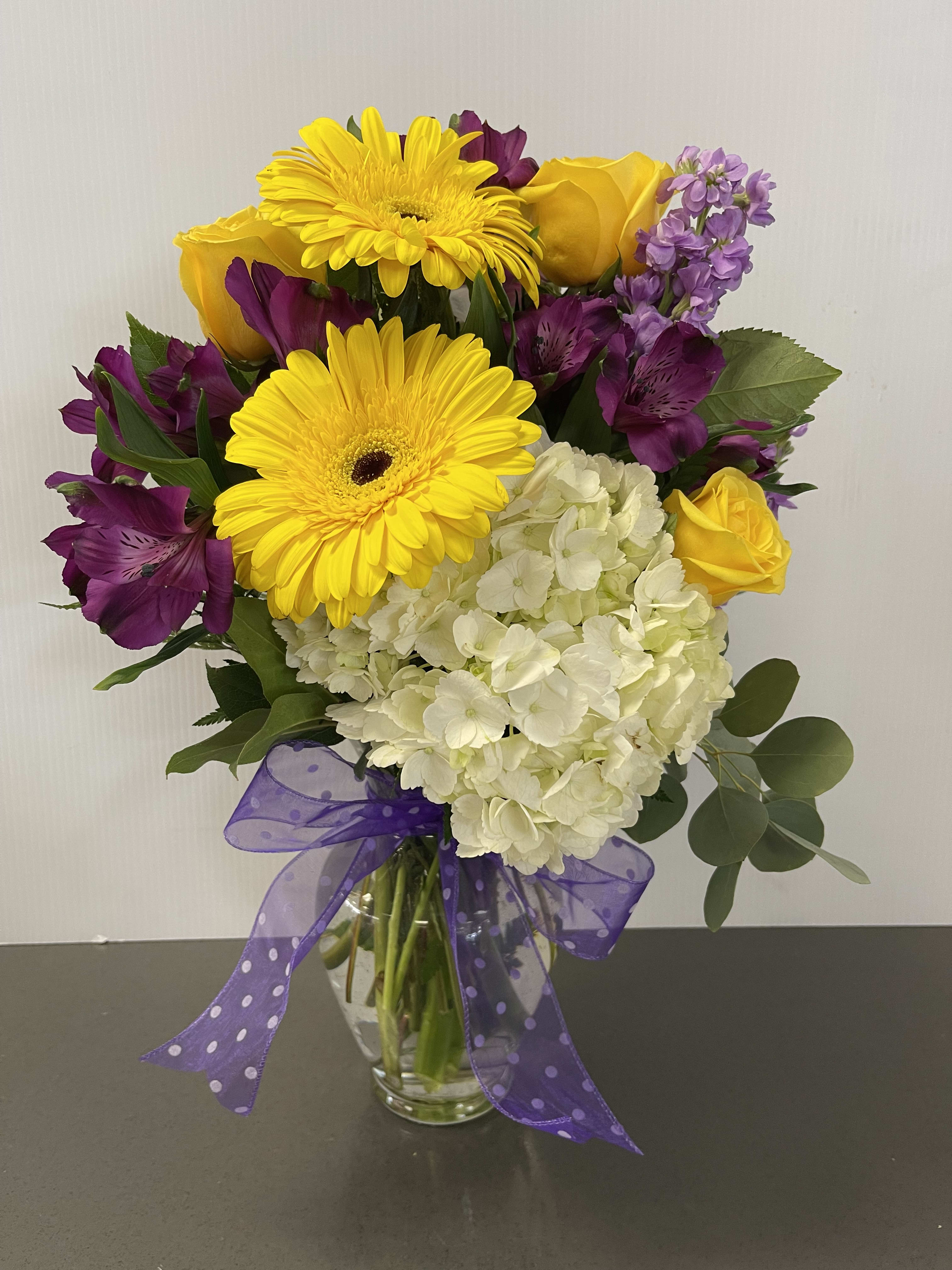 Bouquet of yellow gerbera daisies, roses, purple flowers, and white hydrangea in a glass vase