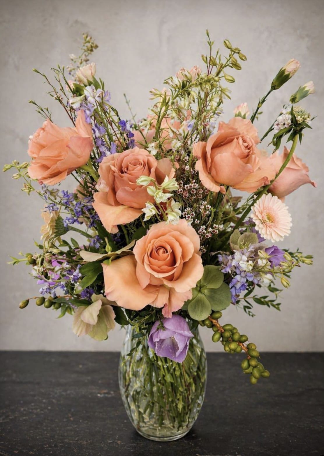 Peach Garden - Soft peach roses take center stage in this garden-style flower arrangement, a Peach Garden bouquet perfect as a birthday bouquet, Mother's Day gift, or an elegant thank-you arrangement. The loose, airy design reads like a fresh-picked posy-tall stems and scattered blooms create an open, natural silhouette that feels both romantic and relaxed for springtime celebrations or everyday gifting.  Mixed daisies and other purple and white accent flowers weave through the roses, adding contrast and delicate texture against fresh greenery. Arranged in a clear glass vase, the composition emphasizes soft peach tones balanced by lavender-hued accents and crisp white blooms for a warm, feminine palette that makes the recipient feel truly seen. The arrangement arrives ready to display in its vase-ideal for sending to someone you want to celebrate, comfort, or simply brighten.