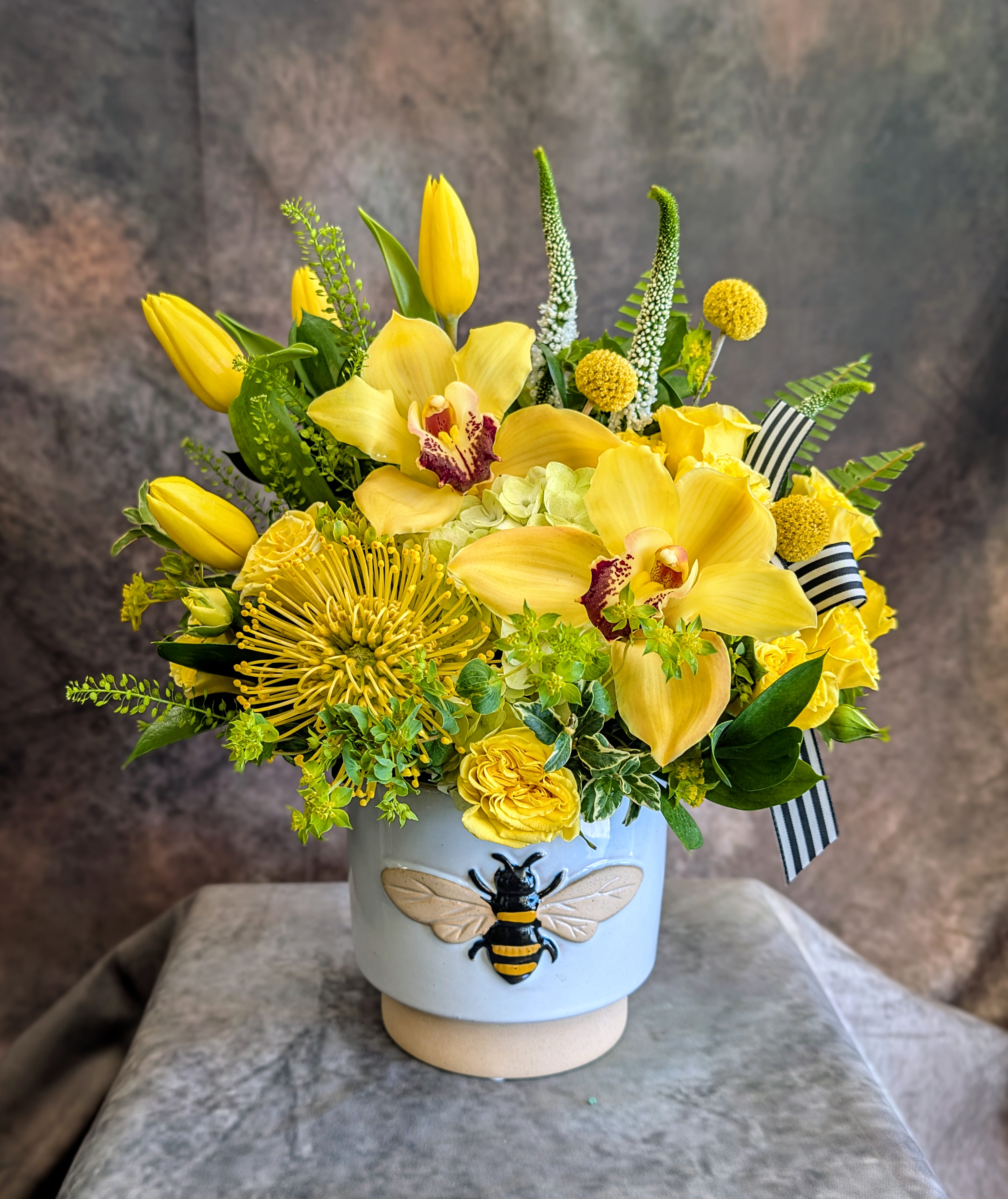 Bumble - Bumble is a joyful burst of golden sunshine, thoughtfully handcrafted at Windmill Floral Expressions in Las Vegas. This cheerful arrangement features luminous yellow cymbidium orchids, vibrant tulips, textured pincushion protea, and playful craspedia, layered with fresh seasonal greens for a lush, garden-inspired feel. Designed in a charming bee-adorned ceramic vessel, Bumble brings a touch of whimsy and warmth to any space—perfect for celebrations, thank-yous, or simply brightening someone’s day.  Each arrangement is custom made with the freshest blooms available, ensuring a one-of-a-kind design every time. Same-day flower delivery is available in Las Vegas and Henderson, making it easy to send a little sunshine when it matters most.