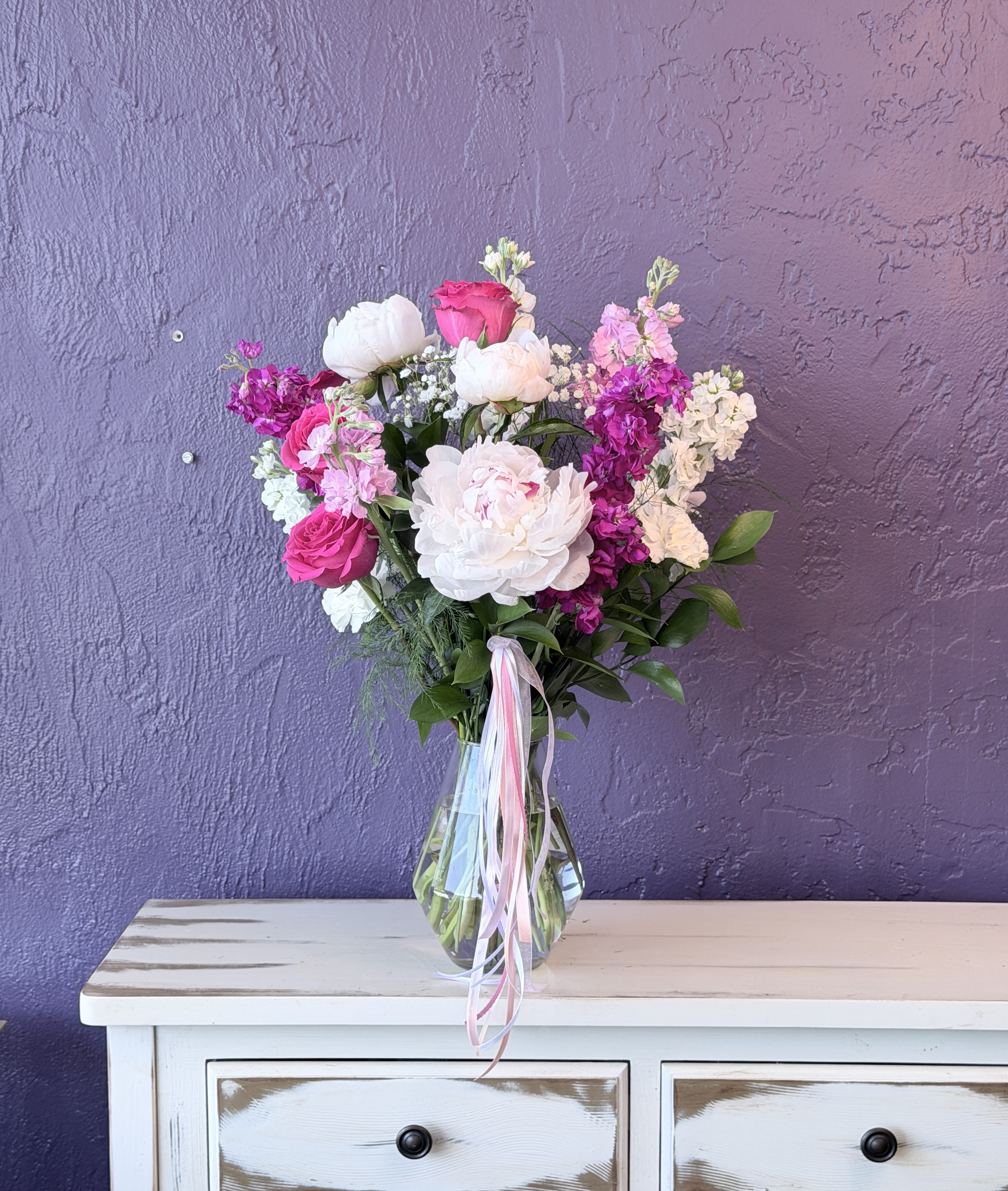 7 Peonies  - Soft magenta peonies and roses create an immediate joyful impact-an upright, garden-style flower arrangement perfect as a birthday bouquet, anniversary gift, or Mother's Day surprise. The mix of pink, white, purple, and green reads feminine and celebratory, and the clear glass vase with pink and white ribbon streamers gives the recipient a ready-to-display arrangement that feels thoughtful and festive.  This design features large peony-like blooms and roses at the center, with clusters of stock, daisies, and other small textured flowers filling the composition for a full, loose look. Dark foliage provides contrast to the bright magenta accents and pure white blossoms, creating depth and movement. The arrangement is presented in a clear glass vase with pink and white ribbon streamers for an elegant finishing touch.  A lovely choice for the person you're celebrating-she'll feel seen and cherished whether it marks a milestone or simply says "I'm thinking of you." Sized to sit on a table or mantel, the vase makes care simple: keep stems trimmed, change water regularly, and enjoy the scent and color while blooms last.