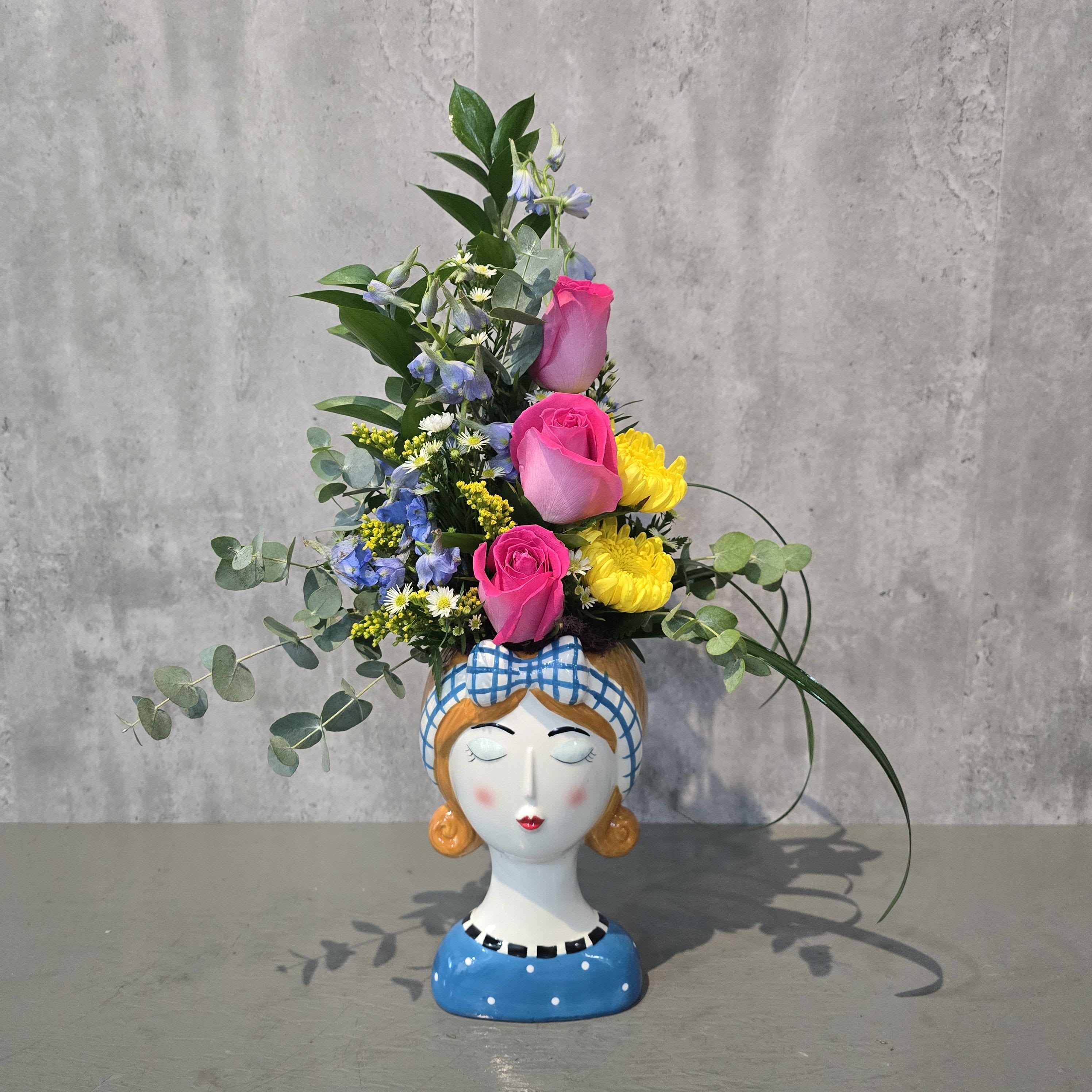 Claire - Claire is a playful, garden-inspired floral design featuring hot pink and light pink roses, bright yellow mums, soft blue delphinium, and delicate white daisy accents, finished with lush eucalyptus greenery.  Arranged in a charming ceramic face vase, this unique piece is perfect for birthdays, thank you gifts, or everyday moments. A beautiful keepsake and a standout option for flower delivery in Albuquerque.