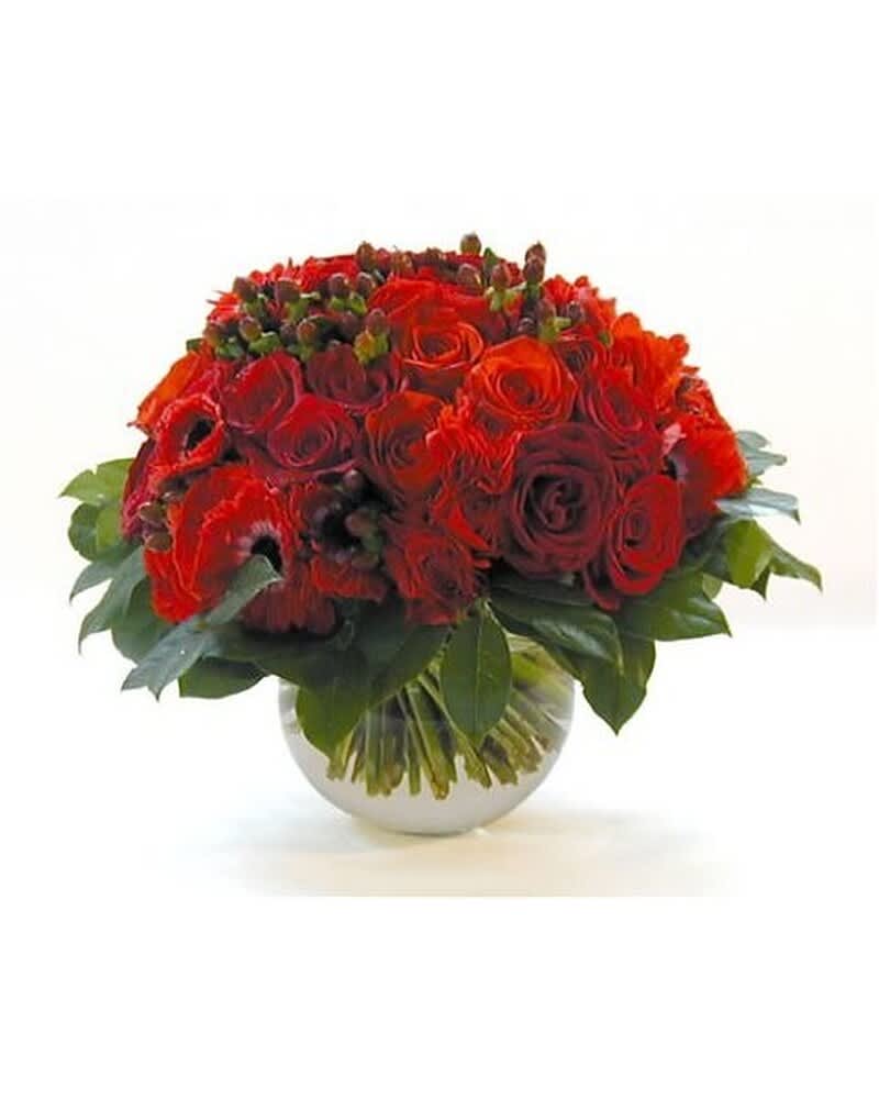 Low round arrangement of red roses in a clear glass bowl vase with a collar of glossy leaves.