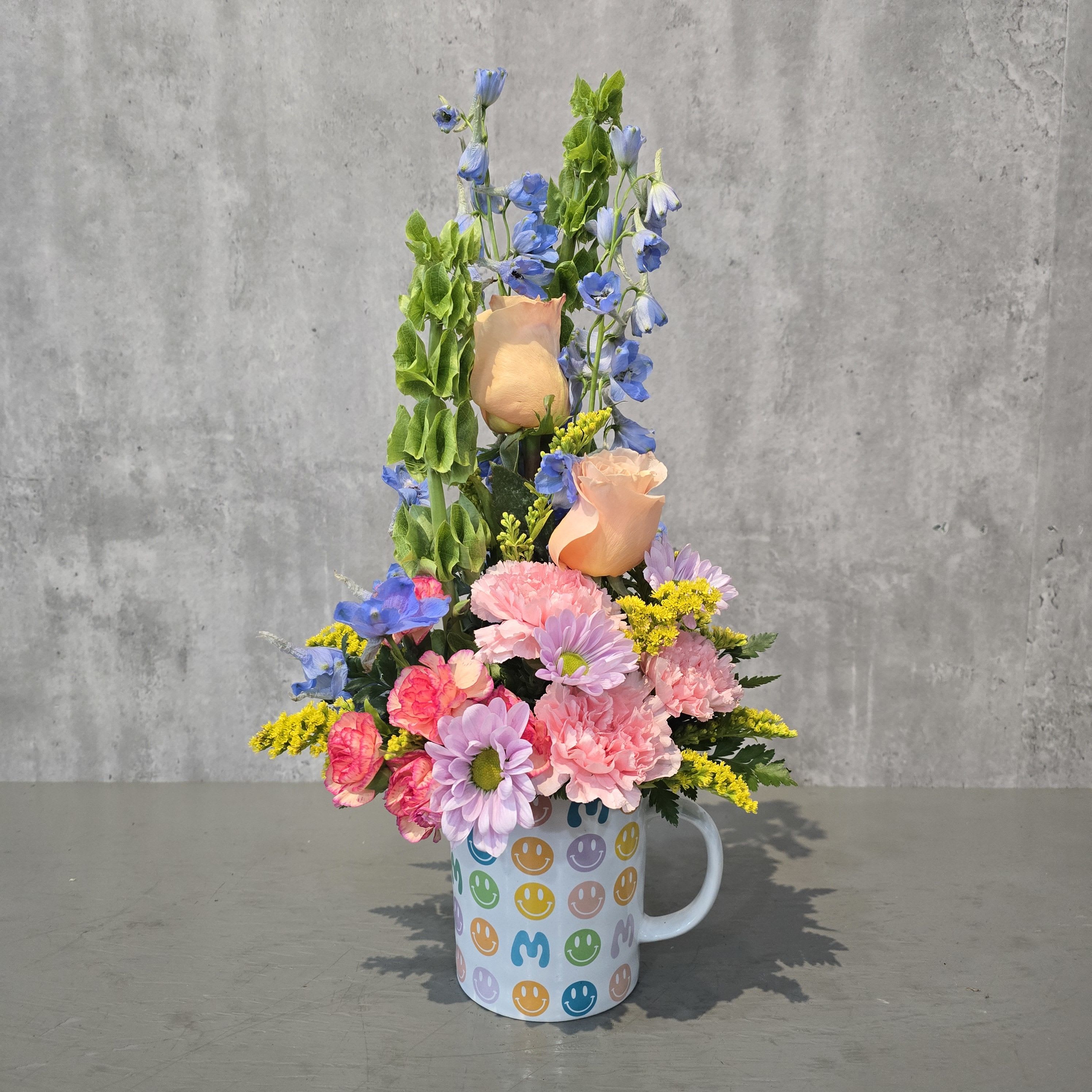 Groovy Mom Garden Mug - A cheerful, retro-inspired design featuring a smiley-face mug filled with peach roses, pink carnations, lavender daisies, blue delphinium, and bright yellow accents, finished with lush greenery.  Perfect for Mother’s Day or birthdays, this playful arrangement doubles as a keepsake mug and is available for flower delivery in Albuquerque.