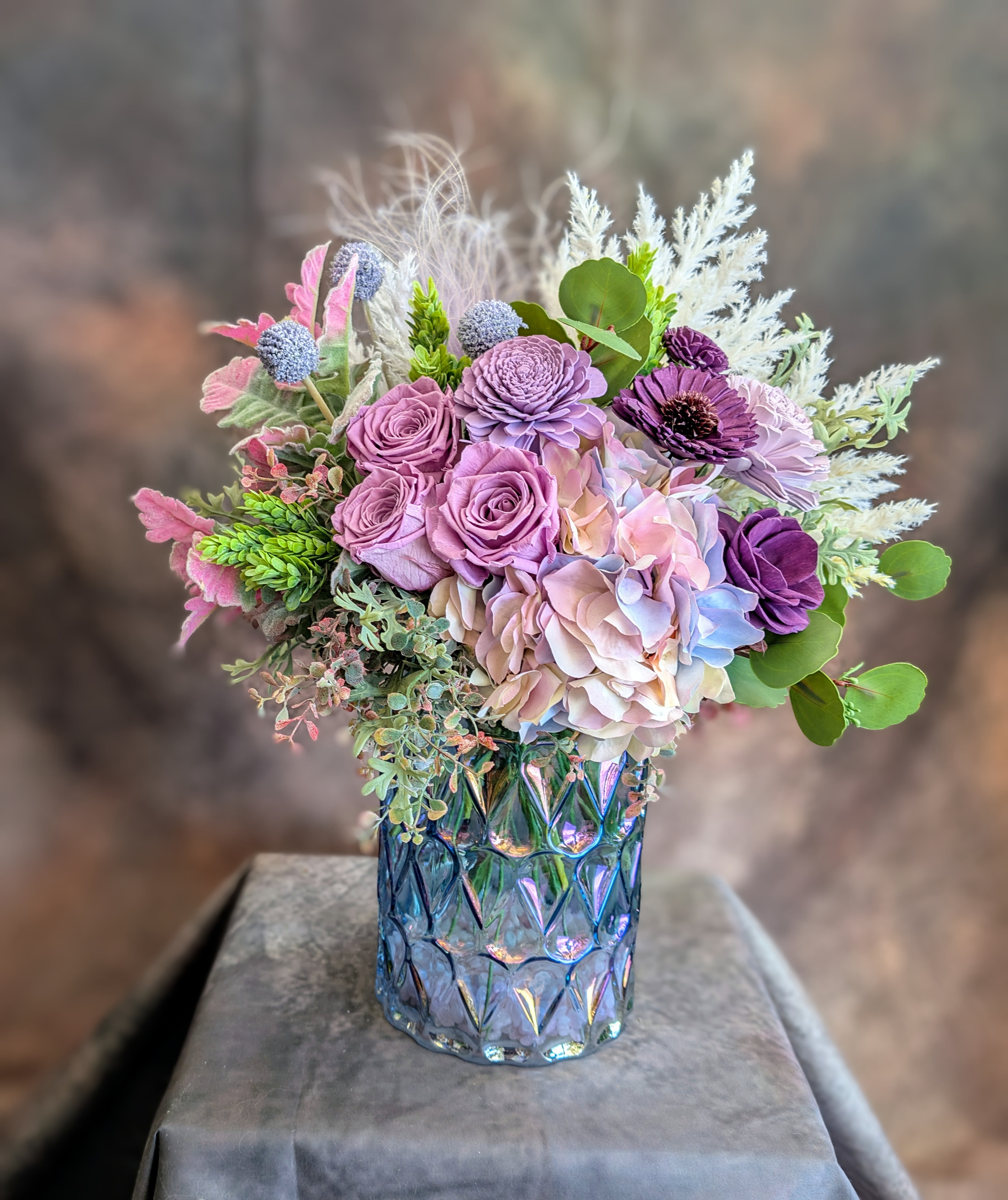Astoria- A Preserved, Sola Wood Flower, And Artificial Permanent Arrangement - Astoria is a softly luxurious arrangement of preserved roses, handcrafted sola wood flowers, and premium artificial botanicals, designed in romantic mauves, dusty pastels, and layered textures for a look that feels both timeless and modern. Featuring real preserved roses paired with intricately crafted sola wood blooms, lifelike ranunculus, and airy hydrangea in shades of lavender, blush, and soft violet, this design is accented with textural elements and sculptural greenery for depth and movement. Set in a luminous iridescent glass vessel, Astoria catches the light beautifully, adding an elevated, glowing finish to any space.  Handcrafted at Windmill Floral Expressions in Las Vegas, each piece is thoughtfully composed using high-quality permanent botanicals, offering long-lasting beauty with no maintenance required. Perfect for home décor, gifting, or keepsakes, Astoria is designed to be enjoyed for seasons to come.  A refined, modern take on classic floral elegance, this arrangement is ideal for birthdays, celebrations, or simply adding a touch of effortless luxury. Available for delivery in Las Vegas and Henderson or curbside pickup by appointment. 