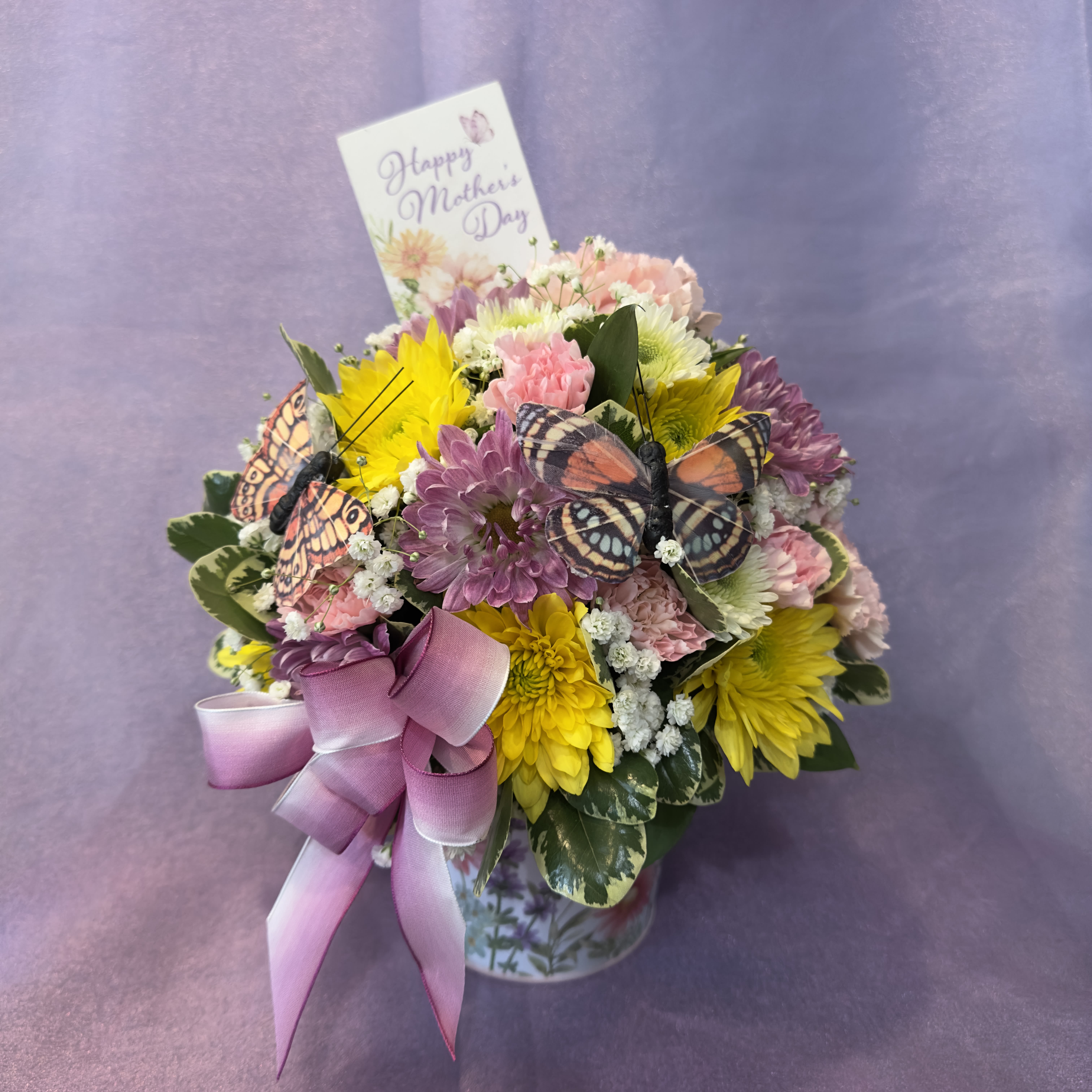 Mother's Day Butterfly - A cheerful Mother’s Day arrangement featuring bright yellow and lavender mums, soft pink carnations, white accent flowers, decorative butterflies, and a pink ribbon bow in a floral keepsake container. Includes a Happy Mother’s Day pick for a sweet seasonal touch. Please keep in mind each arrangement designed might have some color changes based on current supply of flowers.