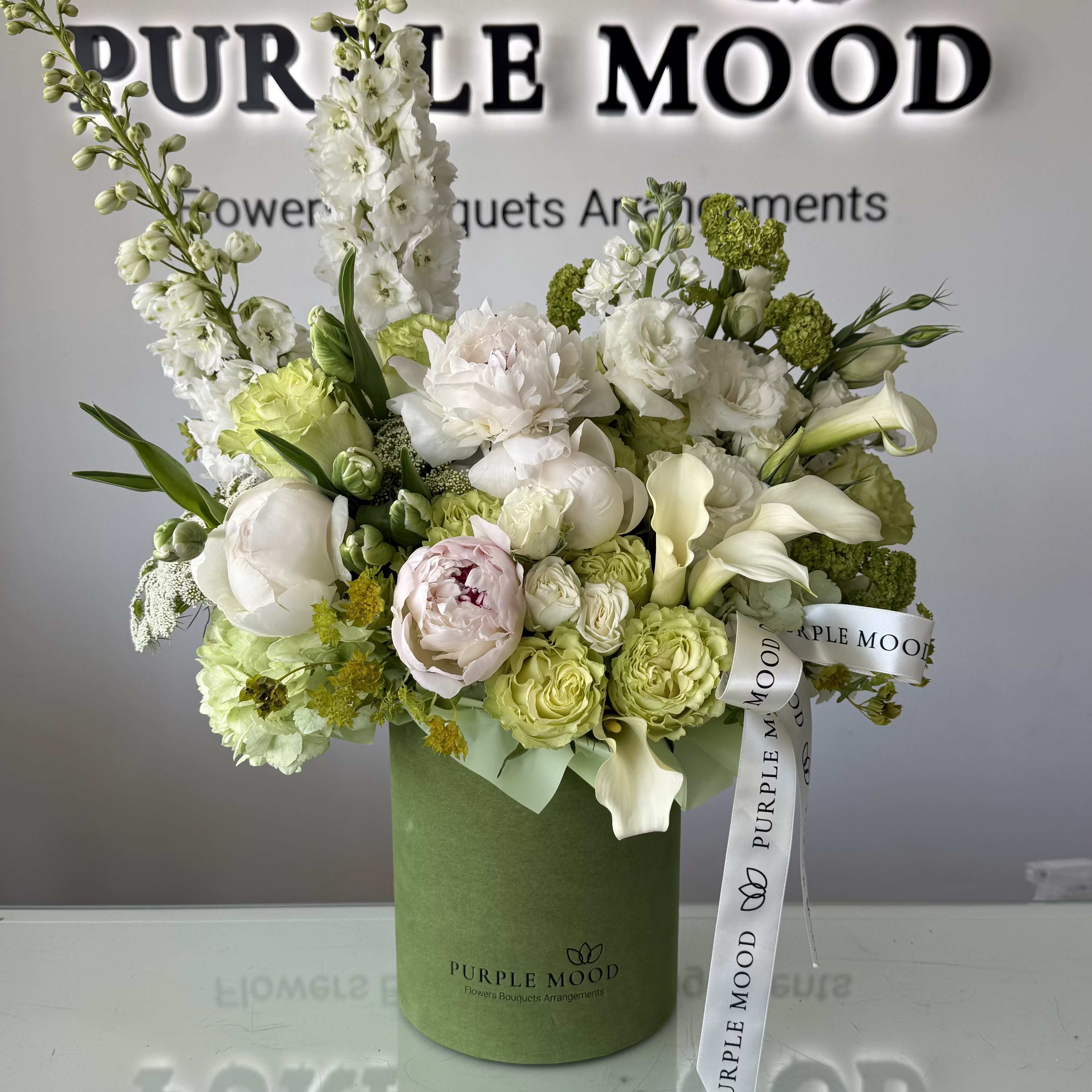 Elegance Box - A fresh, elegant arrangement designed in soft green and ivory tones, this piece blends lush garden textures with a modern, clean look. Featuring premium blooms like peonies, roses, lisianthus, delphinium, and calla lilies, it’s arranged in our signature green hat box for a refined, elevated presentation.  The combination of airy whites and soft greens creates a calming, sophisticated feel—perfect for Mother’s Day, birthdays, or simply bringing a touch of beauty into any space.  Details:  Designed in a hat box (with lid) Arranged in floral foam for long-lasting freshness Includes premium seasonal blooms in white and green palette
