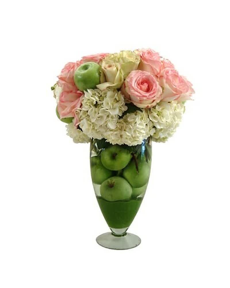Tall glass vase filled with green apples topped with white hydrangeas and pink roses