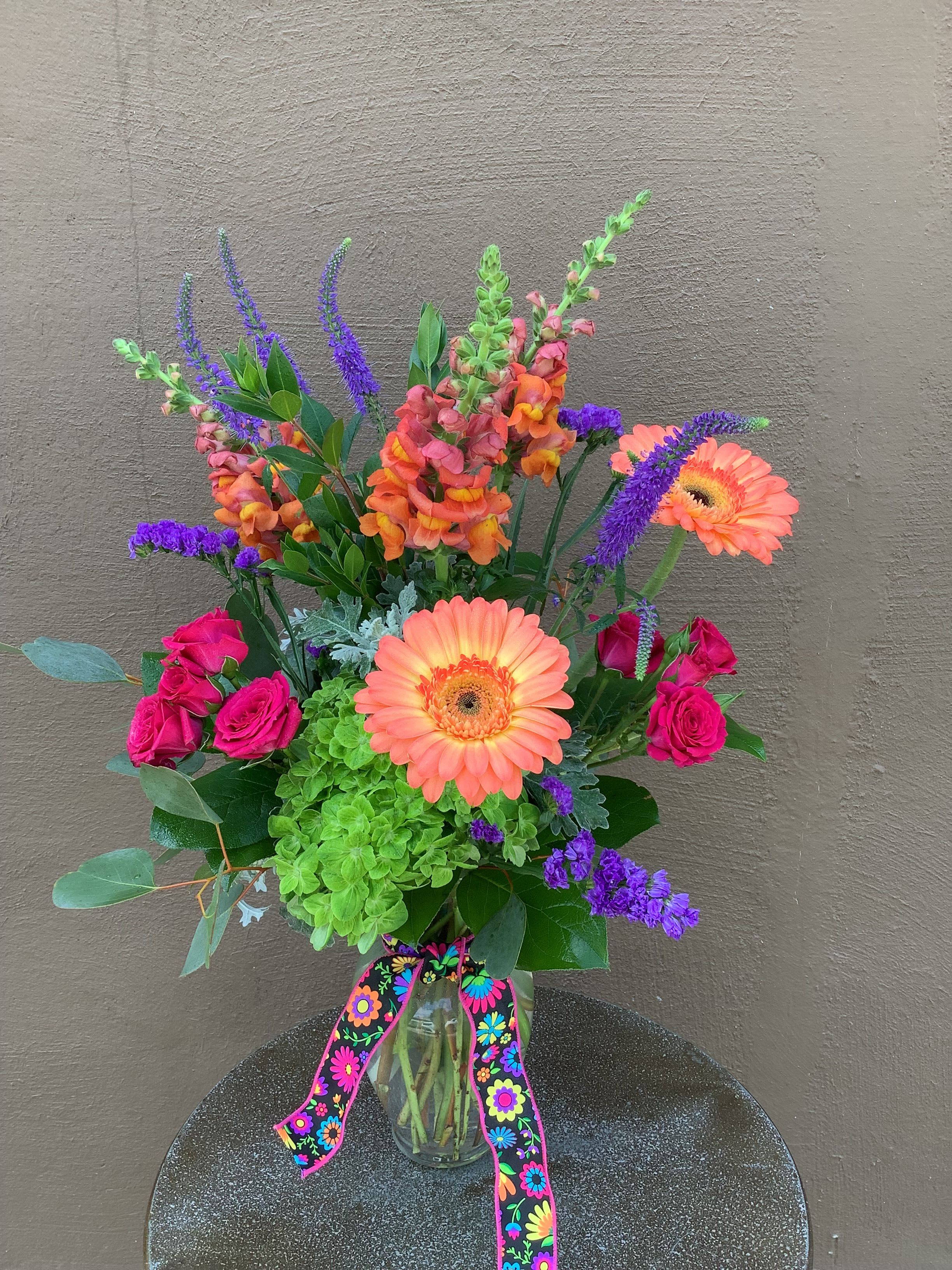 Love to mom - A burst of pink, coral and purple garden-fresh blooms that reads like a joyful bouquet for Mom - perfect as a Mother's Day gift, a birthday surprise, or a bright "just because" to make her smile. This tall, airy flower arrangement sends an uplifting message: she'll feel noticed and celebrated the moment she sees the lively mix reaching upward and outward from the vase.  Hand-arranged in a clear glass vase with a colorful decorative ribbon, the design pairs vivid pink roses and coral-orange gerbera daisies with purple spiky blooms, spray roses and a large green hydrangea cluster for contrast and texture. The composition has a loose, garden-style silhouette that looks especially fresh in spring and summer but suits get-well wishes, apologies, or congrats any time of year. Vase included; stems are styled for a long-lasting display - simply trim stems and refresh the water to keep the arrangement looking its best.