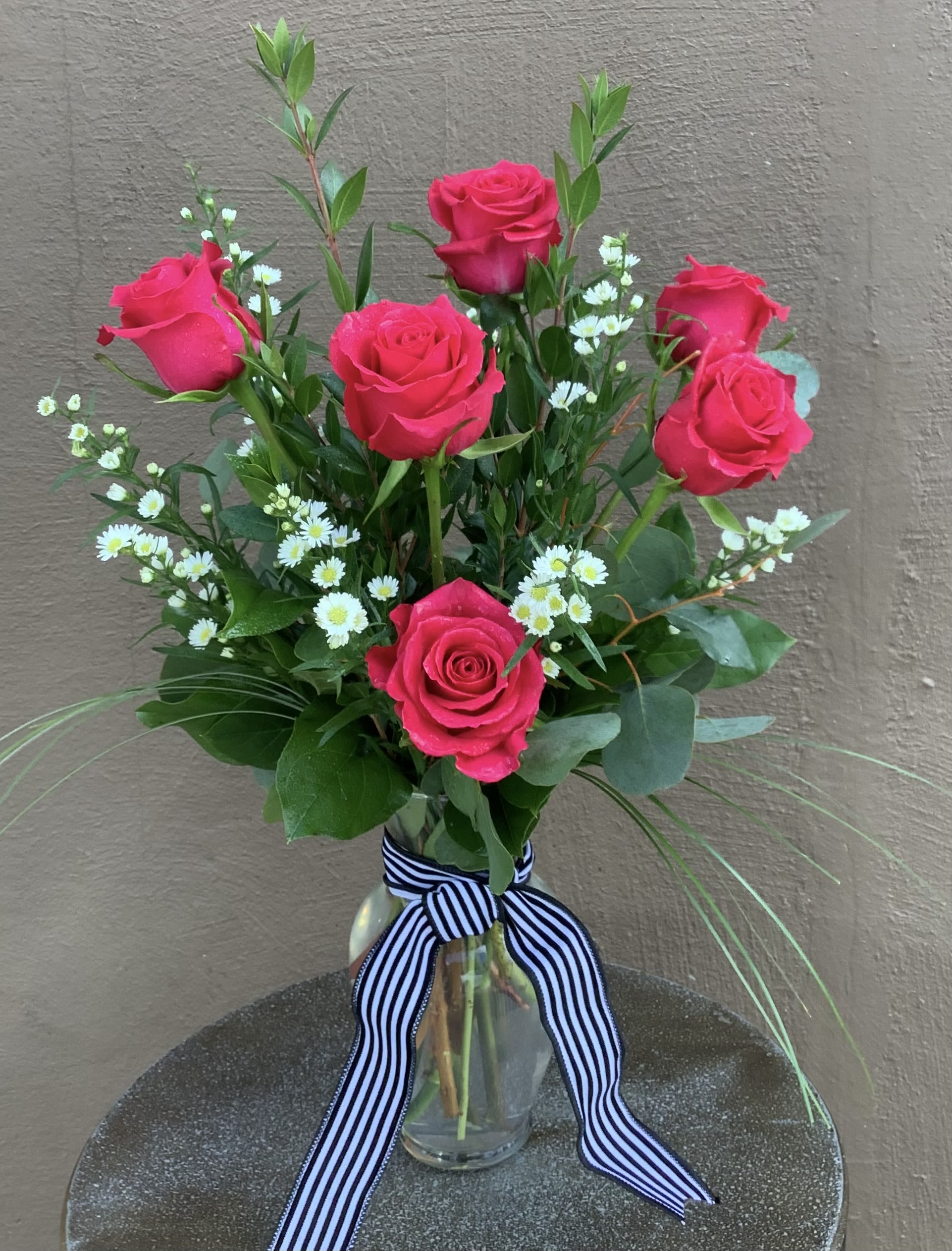 6 long stem roses - various colors available  - Vivid pink roses gathered in a clear glass vase make an immediate, joyful statement-perfect as a birthday bouquet, anniversary roses, or a just-because surprise that says "I'm thinking of you." This six long-stem rose arrangement (available in mixed, pink, white, green, or black tones) pairs classic rose blooms with small aster/daisy-like filler and airy greenery for a loose, garden-style silhouette that feels fresh and effortless-an ideal choice for Mother's Day, a romantic gesture, or a gentle apology.  Each stem is arranged at varied heights for an open, natural look and tied with a crisp black-and-white striped ribbon for clean contrast. The display arrives ready in a clear glass vase, sized to showcase the long stems and keep blooms upright. She'll notice the color, texture, and thoughtful presentation right away-whether you're saying thank you, celebrating love and romance, or brightening someone's day just because. Care: keep water fresh, trim stems every few days, and place away from direct heat to extend vase life.