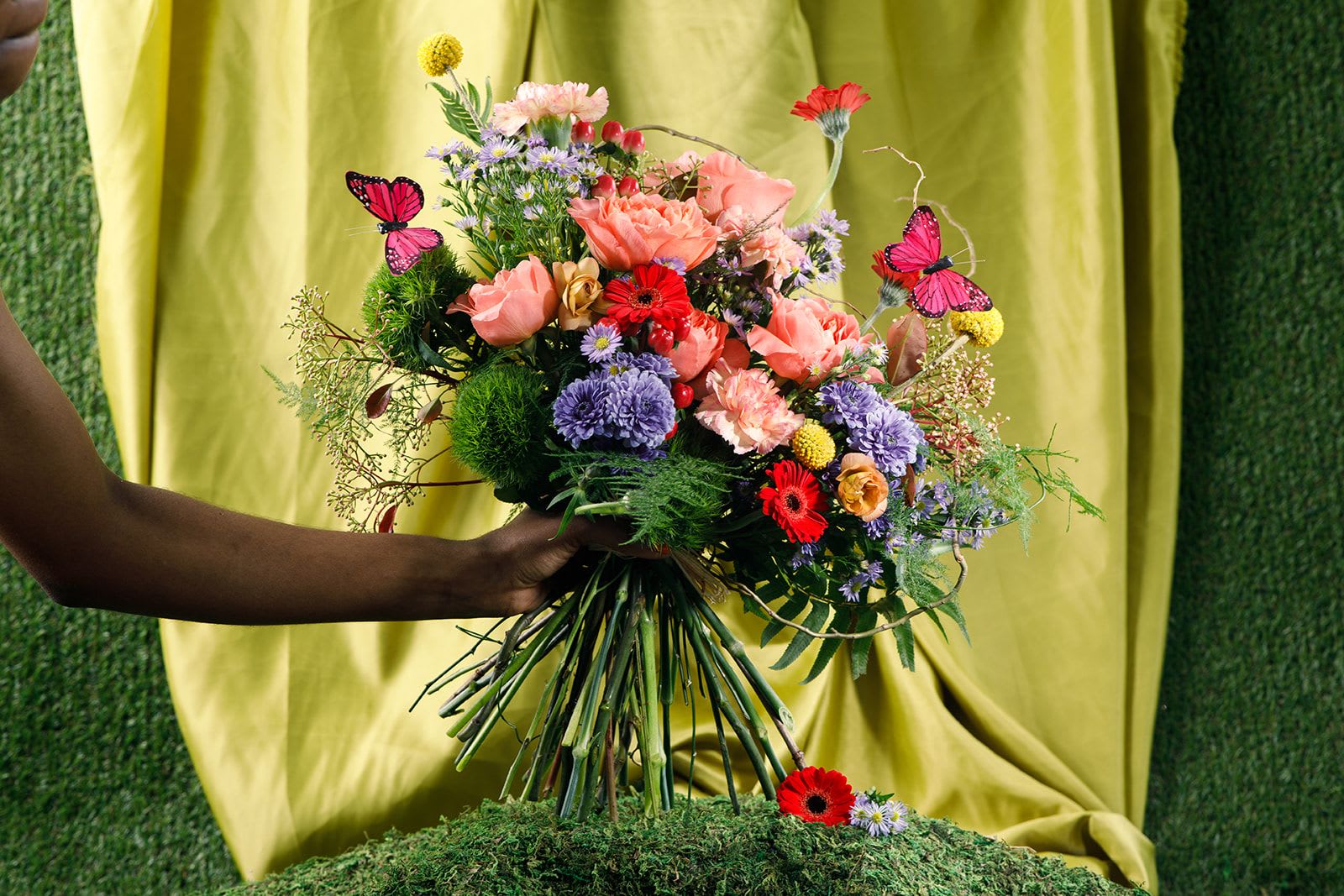 Ladies of the Garden - Ms. DragonFly  - The Intentional Mother  A whimsical hand-tied bouquet that feels like a secret garden come to life - perfect as a Mother's Day bouquet to celebrate a mother who brings color and curiosity to every day. Ladies of the Garden - Ms. DragonFly mixes coral-pink Dragonfly roses, bright red micro gerbera daisies, butterfly ranunculus, and sunny yellow crespida for a lively, playful palette that reads both joyful and whimsical. Decorative butterfly picks and airy greenery flutter through the stems, giving the arrangement a light, garden-inspired motion that echoes the delicacy of a dragonfly.  Give this to the mother who guides with gentle strength and wonder - she'll feel seen, celebrated, and a bit whimsy. To keep the arrangement fresh, trim the stems and refresh the water every two days.