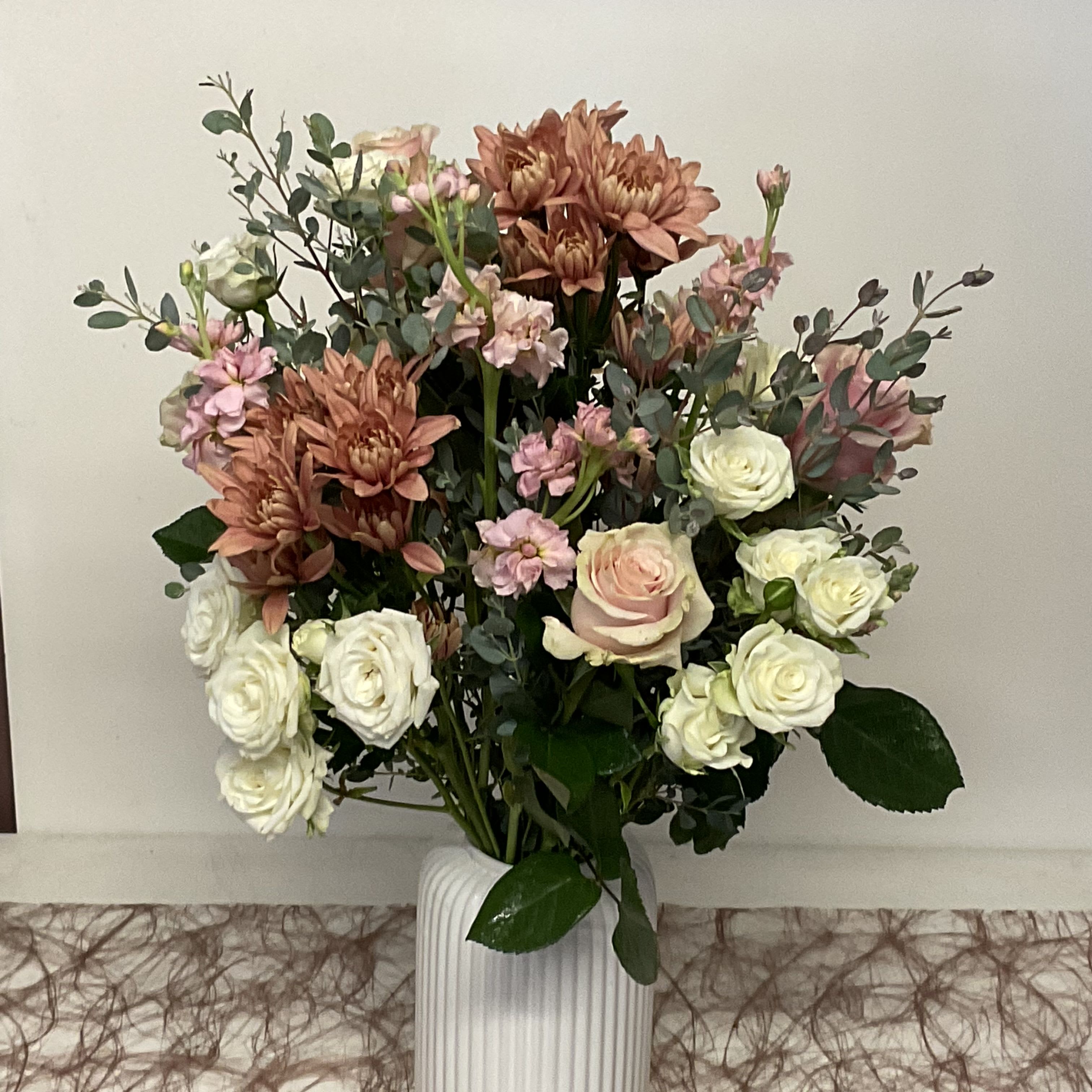 26-127 -  White, pink, peach and green roses, spray roses mums and mini carnations arranged full in a tall white ceramic vase.