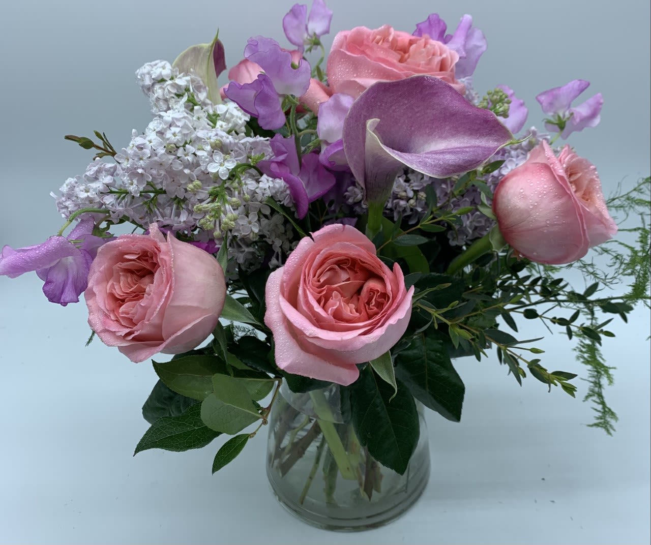 JEANNE'S DAYDREAM $135/165/195 LILAC - This low and lush design includes soft pink Calla Lilies, pink Garden Roses and fragrant Lilac.   A vintage collection of Mother's Day favorites. Pictured at regular price, upgrades include additional fresh blooms.  PS Don't forget to add some chocolates or a fragrant candle!