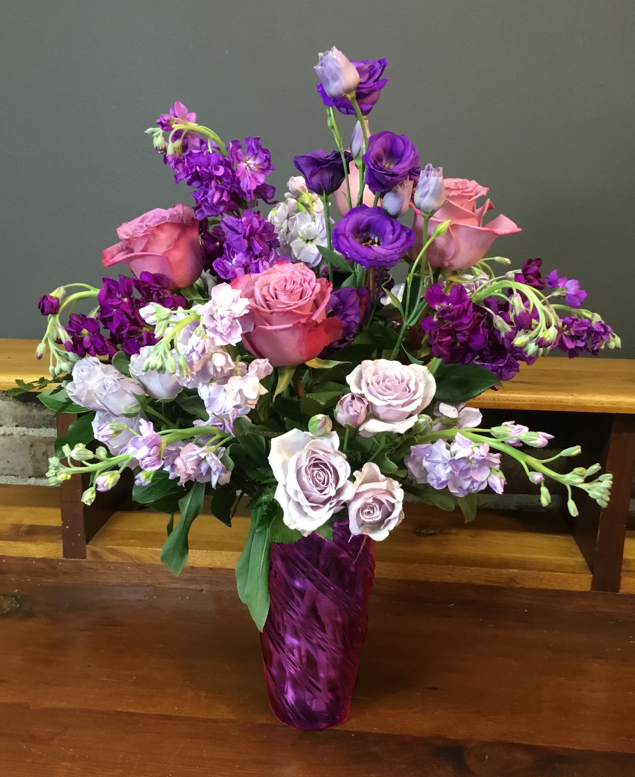 Purple Passion - A lush sweep of rich purples makes this Purple Passion flower arrangement a memorable choice for an anniversary bouquet, birthday surprise, or Mother's Day gift. The rounded, abundant shape and romantic color story-deep violet tones balanced by pale lilac and blush-creates an immediate sense that the person you're thinking of is truly celebrated.  Fresh lisianthus, classic roses, fragrant stock, and delicate spray roses are layered to give texture and movement, with lavender and white lisianthus punctuating the deeper purple blooms. The stems arrive gathered in a tall purple glass vase that reinforces the saturated palette and stands beautifully on a dining table, desk, or mantel. This arrangement is equally fitting for graduations, housewarmings, get-well wishes, or simply saying "thinking of you," and it's sized to make an elegant statement without overwhelming a room.