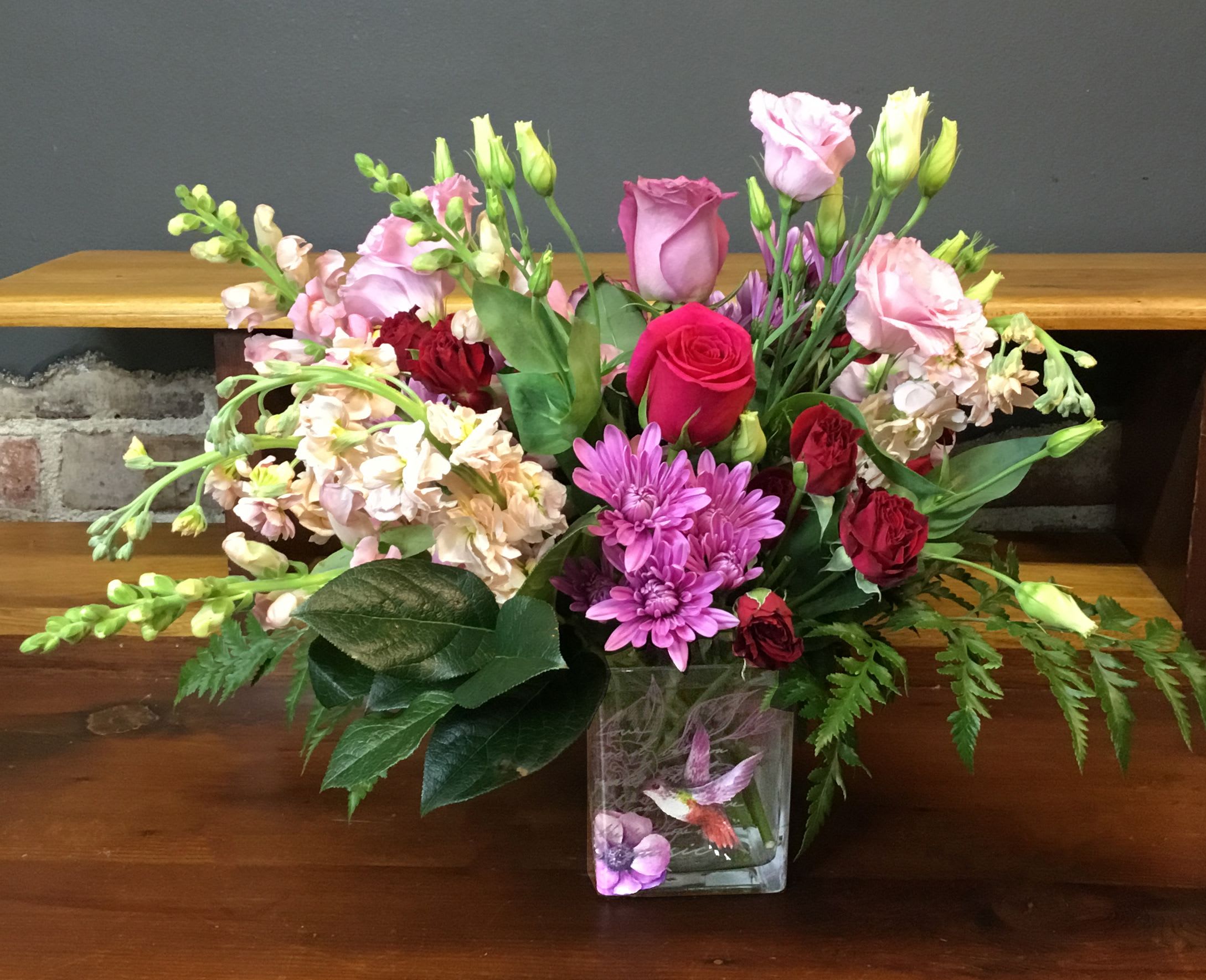 Hummingbird Vase Bouquet - A lush, garden-style bouquet that feels like a personal visit from spring - the Hummingbird Vase Bouquet blends pink and red roses with purple chrysanthemums, pale stock, and delicate pink lisianthus for a romantic, cheerful impact perfect for an anniversary, birthday, or Mother's Day. This flower arrangement communicates love and appreciation in a way that makes the recipient feel truly seen, whether you're celebrating a milestone, saying thank you, or surprising someone "just because."  Arranged full and layered in a clear square glass vase, blooms extend outward for a softly structured, slightly formal silhouette that still reads fresh and garden-fresh. Colors range from soft pastels to saturated red, creating visual depth and texture across roses, spray roses, chrysanthemums, lisianthus, and stock. The compact square vase keeps this bouquet suitable for tabletops, office desks, or bedside tables; it arrives ready to display and bring an uplifting fragrance and cheerful presence to the room.