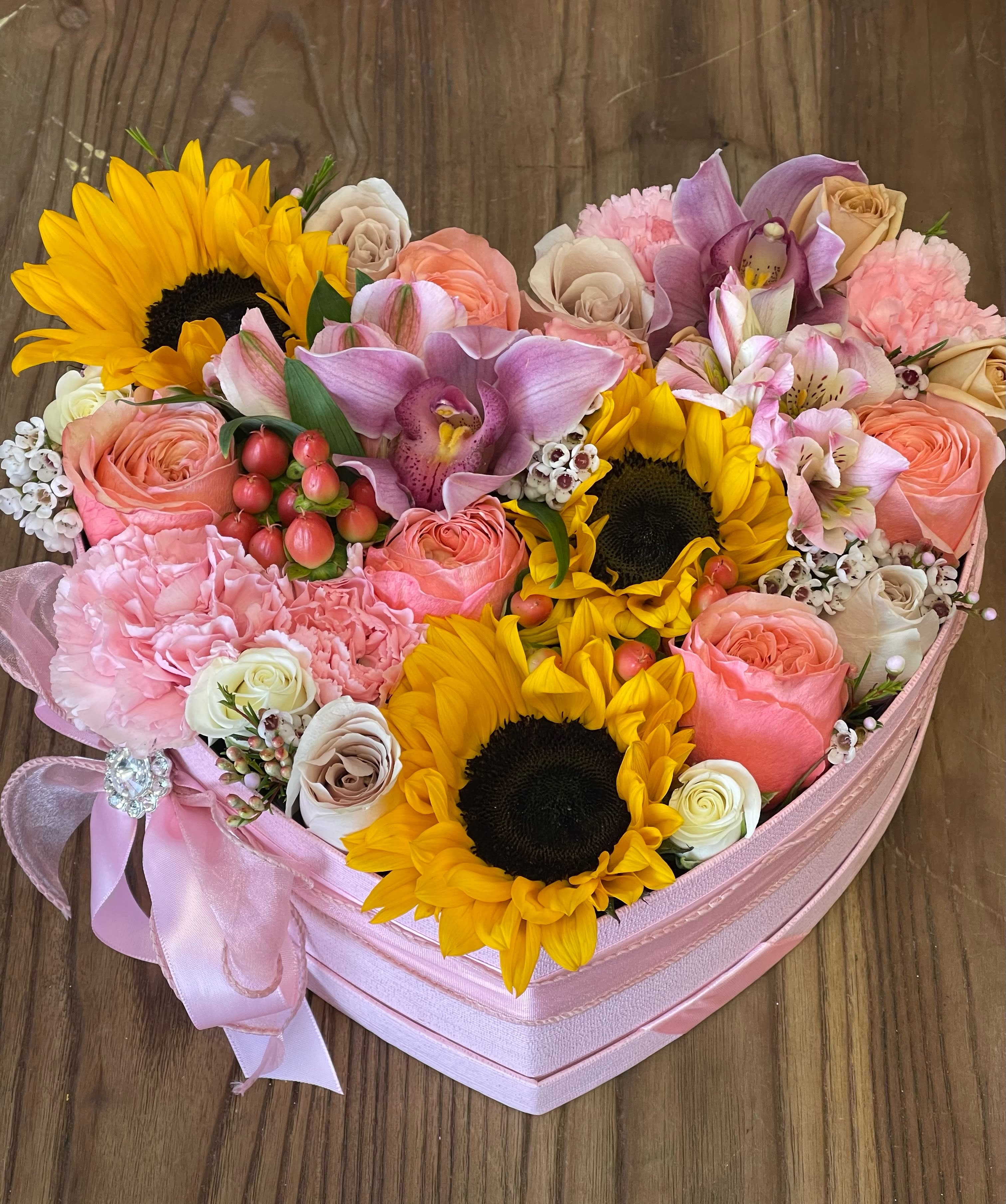 Heart-shaped pink box filled with sunflowers, roses, orchids, and other mixed flowers.