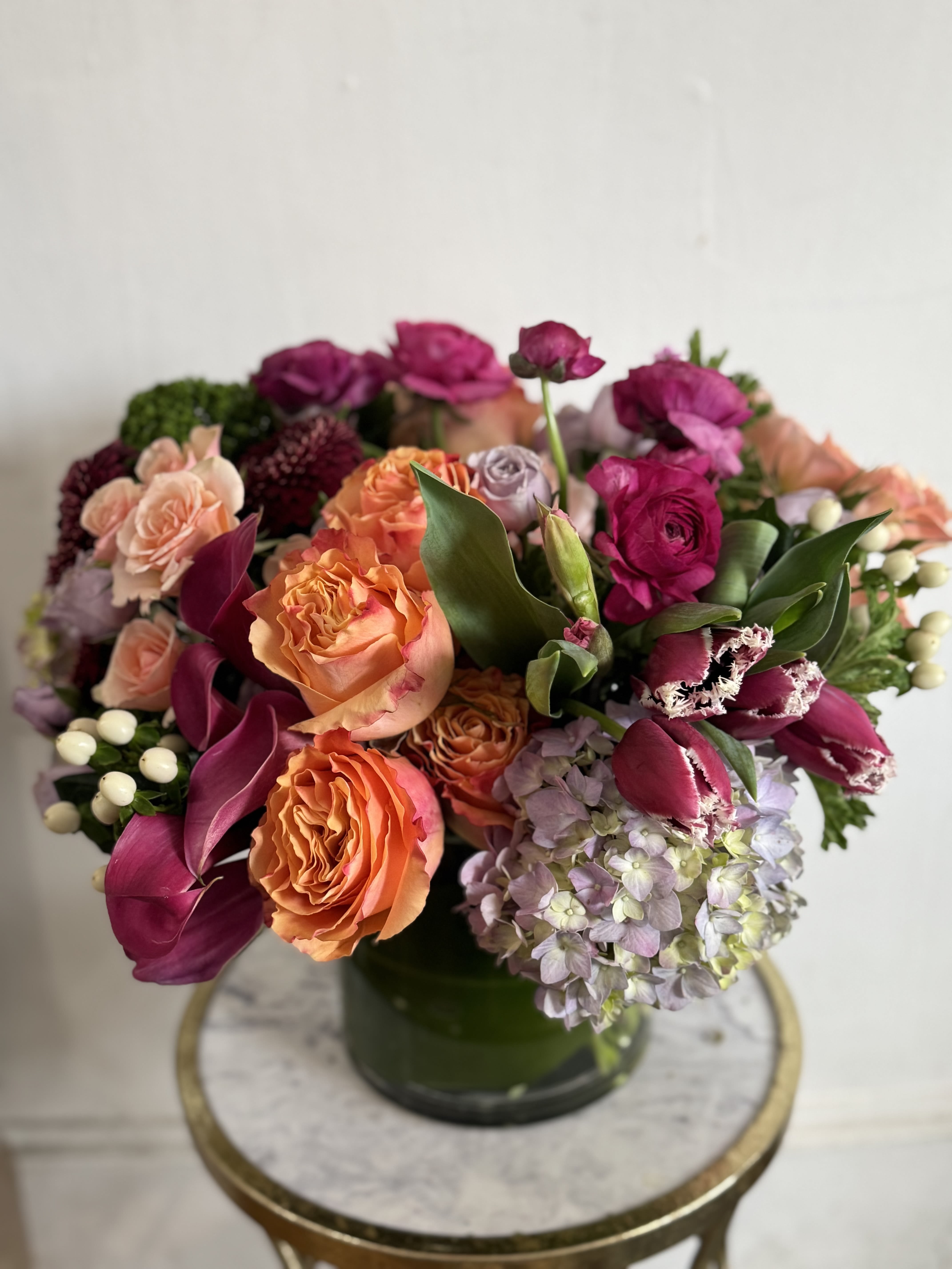 Momosa  - A lush, jewel-toned bouquet that feels like a celebration in a vase-perfect as a Mother's Day gift, a "just because" surprise, or a thoughtful sympathy arrangement. A glass vase holds a dense, tightly packed mix of pink, orange, purple and white blooms so the person you're thinking of will feel wrapped in color and texture the moment it arrives.  Warm coral and orange roses sit against cool lavender hydrangea and deep purple tulips, while magenta and pale pink accents bring brightness; small berry-like accents and graceful calla shapes add movement around the edges.   For the recipient, this bouquet says comfort, celebration, and care-whether you're marking a milestone, offering condolences, or brightening an ordinary day. Arrives ready to display in the included glass vase; replace water and trim stems every few days to extend bloom life.