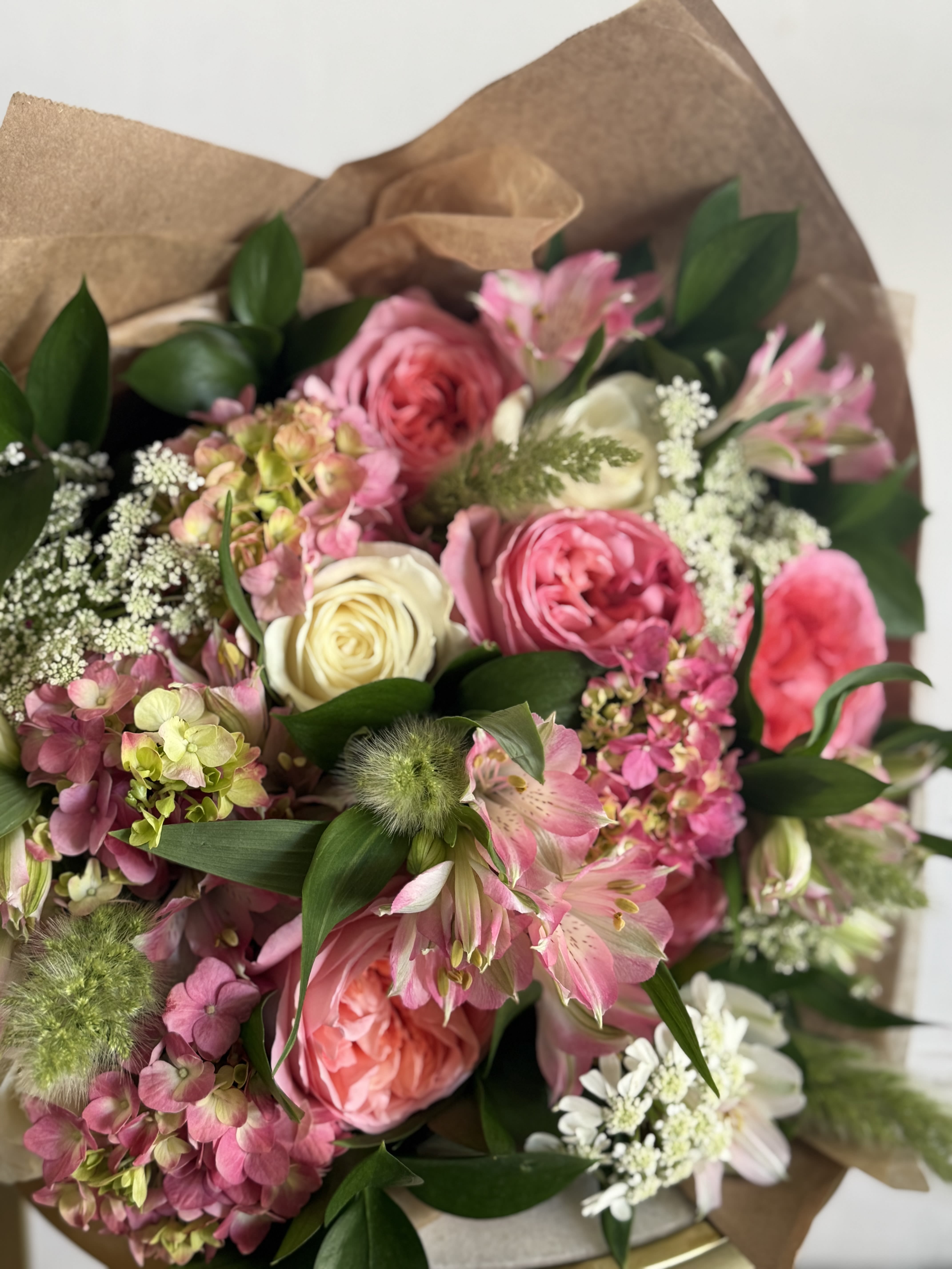 Cream + Pink  DESIGNERS CHOICE  - Soft pink and berry tones mingle with creamy whites in a hand-tied bouquet. Paper wrapped. Designers Choice   This generous arrangement layers pink, white and green. Sized to read abundant and garden-fresh, it arrives loose and tactile - simple care (fresh water and occasional stem trims) keeps the blooms looking their best. Orders are designer's choice to guarantee the freshest premium flowers for Mother's Day, funerals, thank-you gestures, or everyday celebrations.