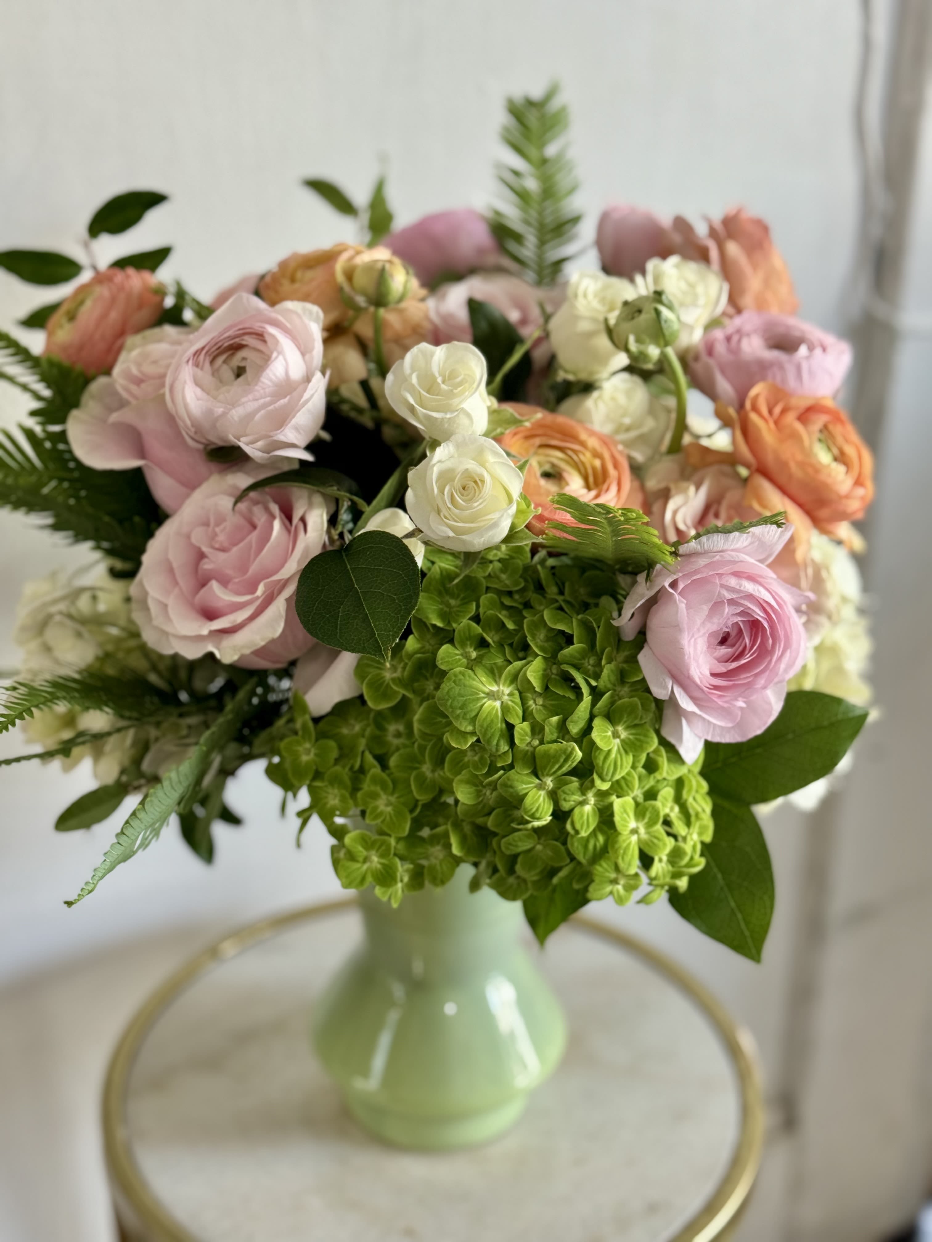 SpringFling  - A lively cluster of soft pink roses, peach ranunculus, and airy white accents crowns a bright green hydrangea centerpiece-an arrangement made to make someone feel delighted and noticed. This spring bouquet is perfect as a birthday bouquet, anniversary flowers, or a Mother's Day gift: the pastel palette reads romantic and fresh, and the garden-style rounded shape feels intentionally effortless, like a picked-from-the-plot surprise.  The design arrives ready-to-display in the vase shown; its compact, rounded profile works well on a bedside table, dining table, or office desk. Choose this for the person you want to make feel truly celebrated-soft, fragrant, and unmistakably spring.