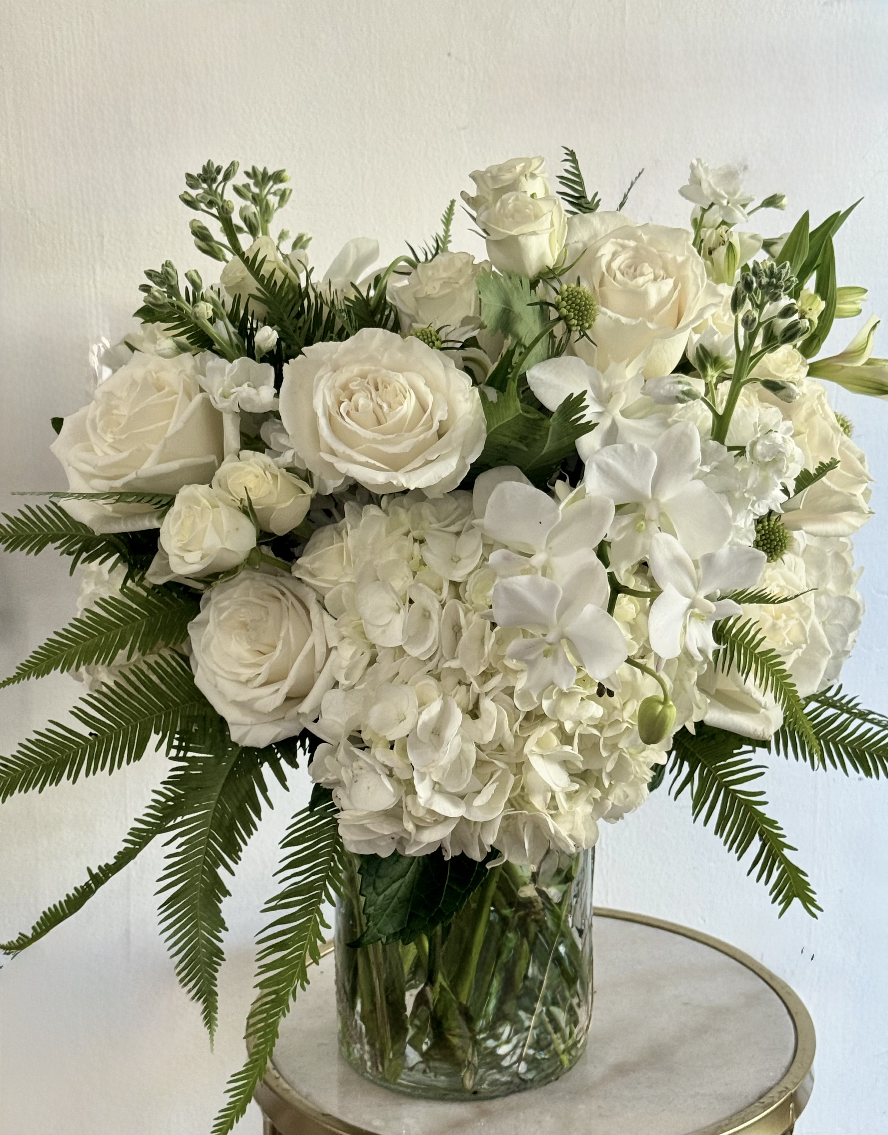 Irreplaceable  - A serene cluster of white hydrangeas and roses, punctuated by delicate white orchids, creates an elegant arrangement that feels quietly consoling and refined. Perfect as a Mother's Day gesture, or a calming get-well gift, this all-white bouquet conveys grace and steadiness-the kind of arrangement that makes the person you're thinking of feel seen and comforted.  The composition is full and rounded, arranged low in a clear glass vase so the blooms read as a single, sculpted mass. Small green textural accents and fern fronds fan gently from the center, adding soft movement and an airy frame to the dense white flowers. Sized to sit on a table or ceremony pedestal, it arrives ready to display-no extra arranging required-and offers long-lasting blooms when refreshed with clean water every few days.
