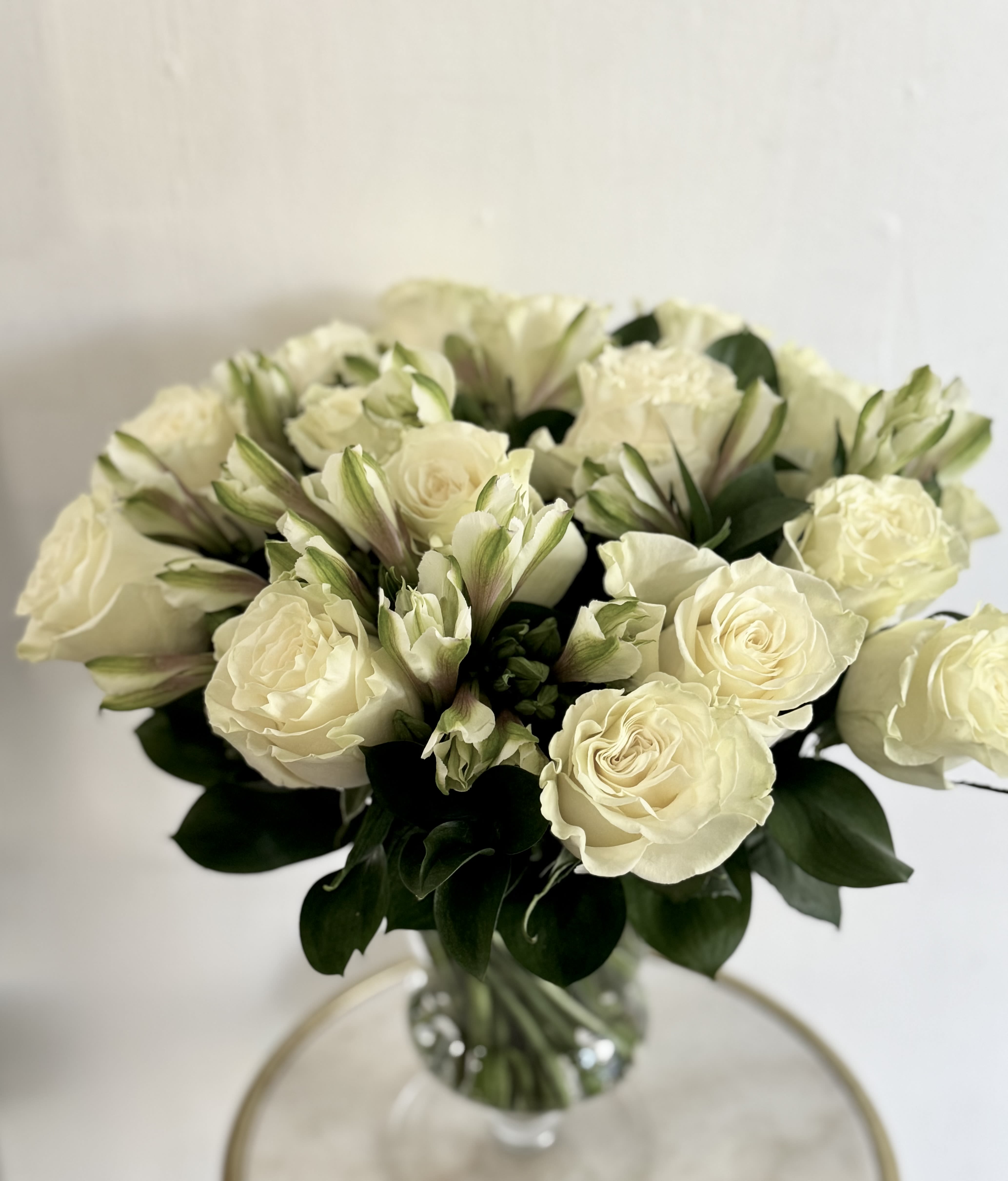 Divine  - Cream‑white roses and delicate white filler form a serene, rounded bouquet that feels quietly elegant-perfect as a sympathy or funeral arrangement, a thoughtful Mother's Day gift,  it also works beautifully as a thank-you or congrats gift when you want to keep the message tasteful and timeless. The soft monochrome palette and subtle green accents read as calming and sincere, the kind of arrangement that lets the person you're honoring feel comforted and truly seen.  Arranged compactly in a clear glass vase, the flowers present a clean, understated silhouette that suits formal occasions or simple "thinking of you" gestures.  The vase keeps stems neatly contained for easy display-refresh water every 2-3 days and trim stems slightly to extend vase life.