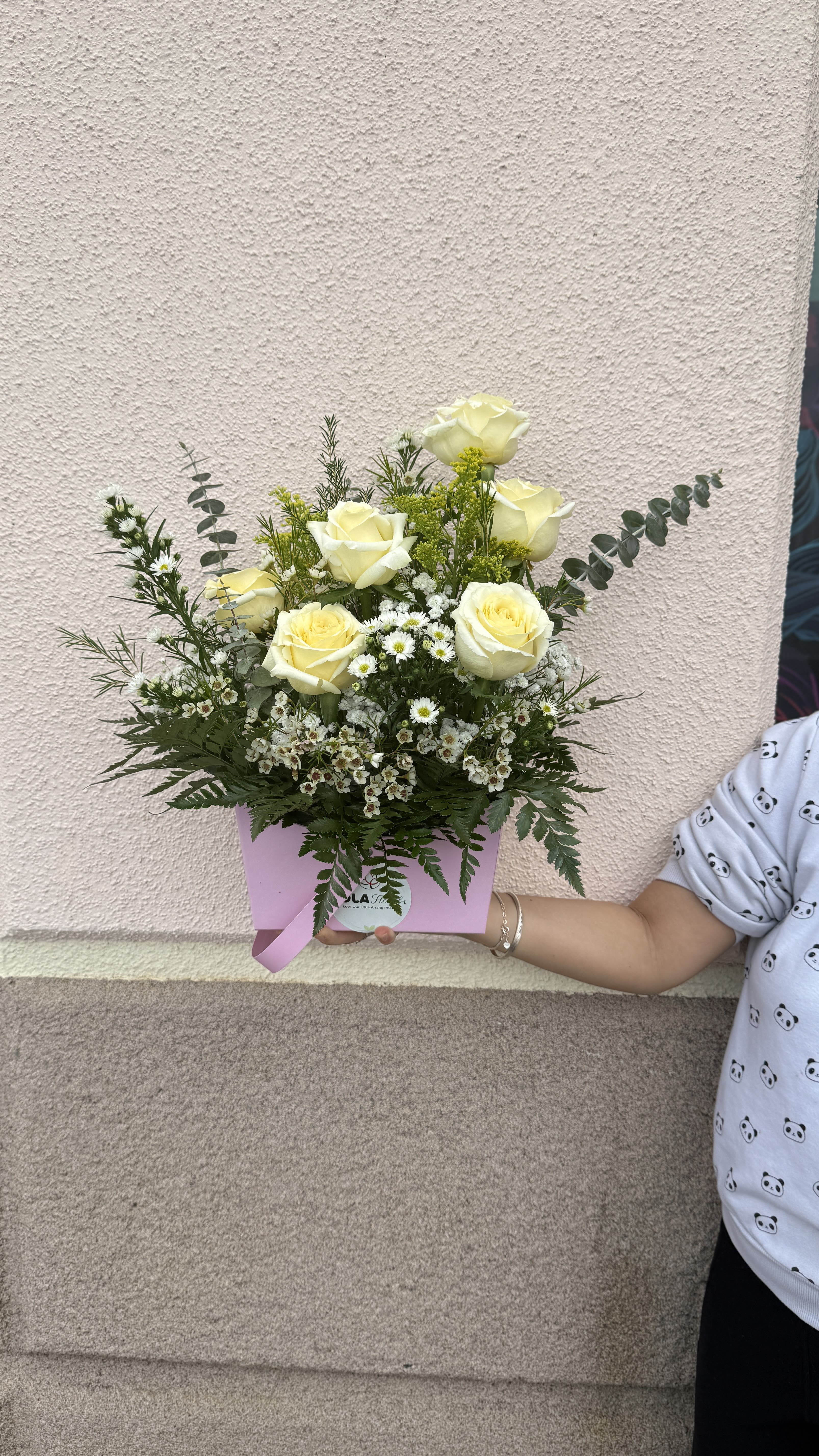 Bloom Box 02  - Soft pale yellow roses clustered above a low pink box create an instant smile in this flower arrangement - perfect as a birthday bouquet, a springtime thank-you, or a cheerful thinking-of-you gift. The round, hand-tied look and delicate white daisy-like fillers give the recipient a gentle, uplifting impression; it reads as thoughtful and feminine, the kind of bouquet that makes someone feel genuinely seen.  This compact Bloom Box 02 arrangement pairs light yellow roses with small white daisy-style blooms and airy green foliage for a full, rounded silhouette. Stems are arranged low in a pink cardboard box tied with a pink ribbon and finished with a round logo sticker, making the presentation feel ready to hand over without extra wrapping. The overall palette of white, yellow, pink, and green keeps the design fresh and versatile for spring celebrations or casual gift-giving.  Sized to sit comfortably on a bedside table or desk, this arrangement is low and lush - easy to display and enjoy. To keep blooms looking their best, replace the floral foam water or refresh stems in clean water if needed; it's designed for immediate gifting and visual impact straight out of the box.
