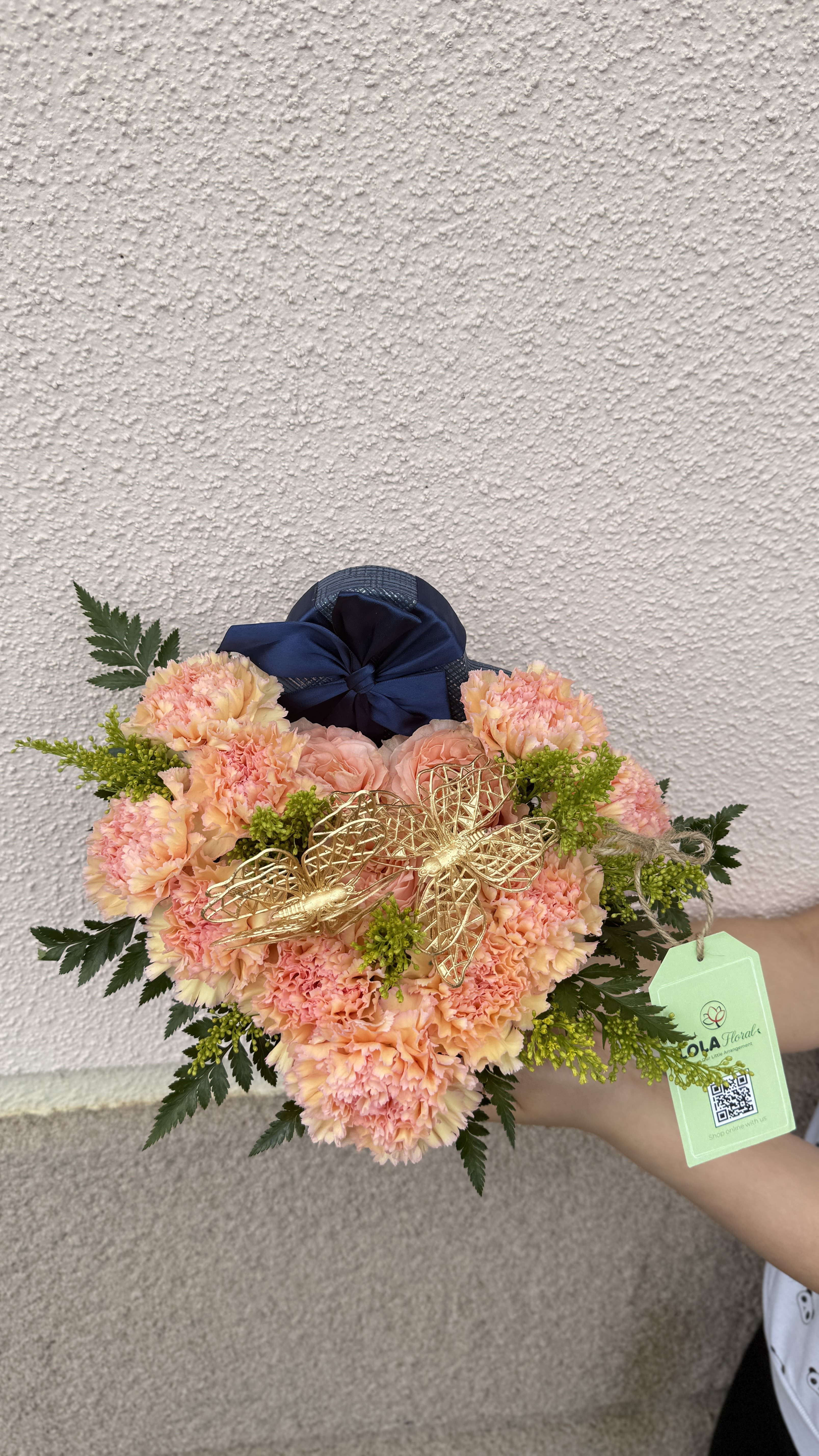 Heart shape box - Peach and pink carnations clustered around soft blush roses create a cheerful, romantic bouquet perfect for a birthday bouquet or a just-because gift. The compact, hand-held shape feels intimate - the kind of arrangement that makes the person you're thinking of feel truly seen and celebrated.  Bright yellow-green filler accents and dark, fern-like foliage frame the rounded composition, while a large navy ribbon, a gold butterfly ornament, and a floral tag give it a polished, gift-ready finish. Compact and handcrafted in appearance, this bouquet pairs playful color (pink, peach, yellow, touches of blue) with classic texture from carnations and roses - ideal for sending warm wishes, congratulations, or a thoughtful anniversary surprise.