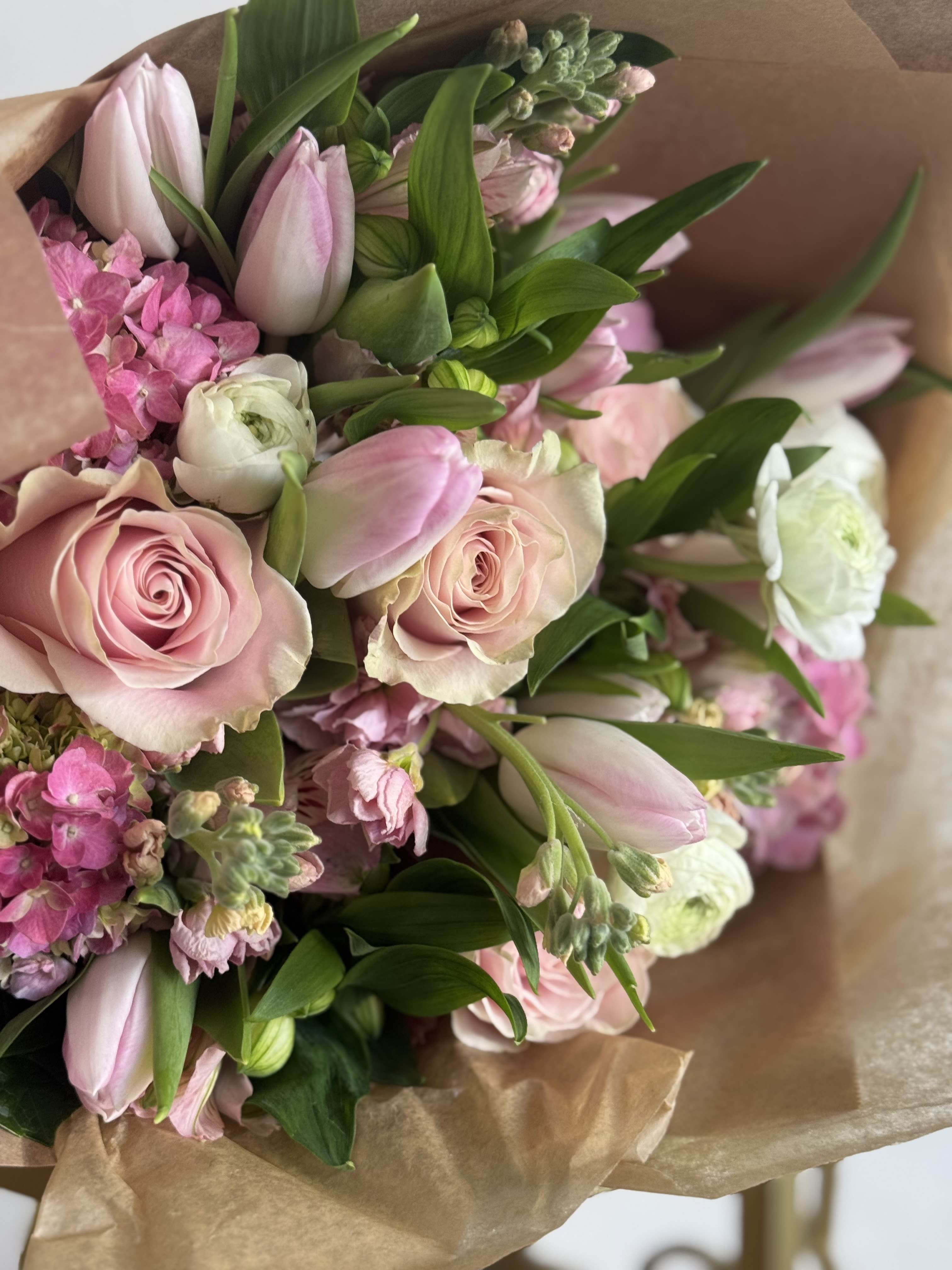 Pink +White DESIGNERS CHOICE - The gentle mix of pinks, whites, and fresh green foliage gives the arrangement an airy, romantic feel that will make the person you're thinking of feel truly seen and cherished. Hand-tied bouquet. Paper wrapped. Designers Choice   This generous arrangement layers pink, white and green. Sized to read abundant and garden-fresh, it arrives loose and tactile - simple care (fresh water and occasional stem trims) keeps the blooms looking their best. Orders are designer's choice to guarantee the freshest premium flowers for Mother's Day, funerals, thank-you gestures, or everyday celebrations.