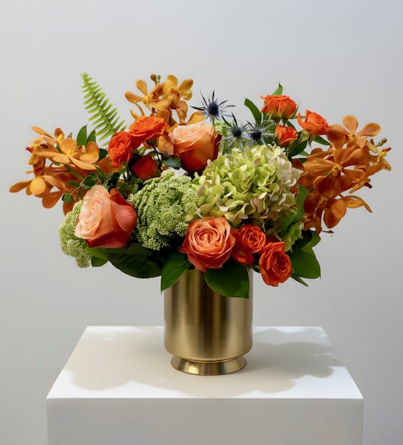 Peaches and Cream - An elegant arrangement in a gold vase featuring vibrant mokara orchids, lush garden roses, and full hydrangeas with complementary blooms. Soft peach tones and creamy neutrals create a warm, refined look—perfect for any occasion.