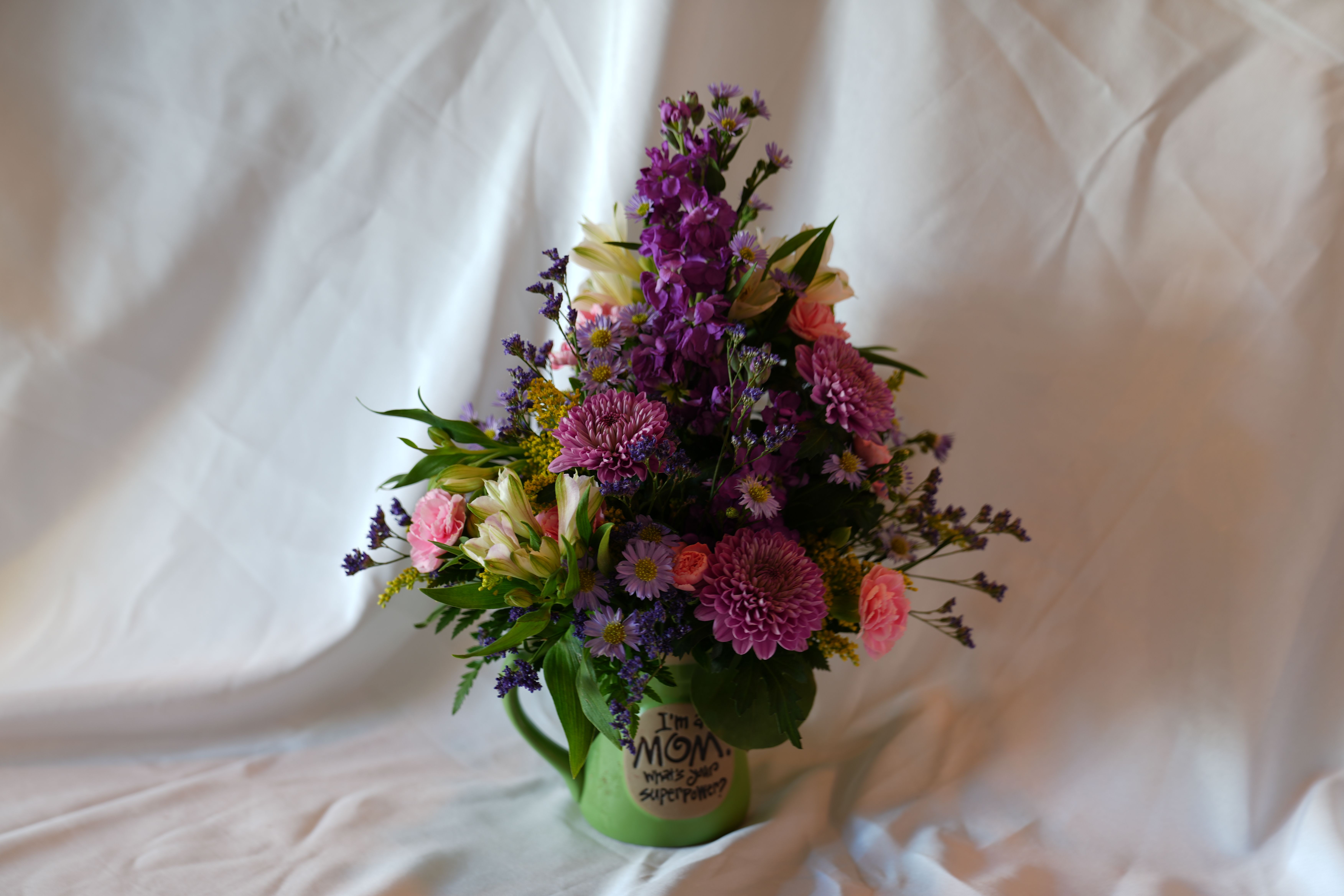 Mom SuperPower - A bright, garden-fresh composition that fits perfectly in a ceramic mug, this flower arrangement is a cheerful Mother's Day gift that will make her feel genuinely celebrated. The compact yet layered silhouette combines purple, pink, white, and yellow blooms to create an upbeat, affectionate impression; it reads like a little bouquet of sunlight and laughter designed for the person you're thinking of.