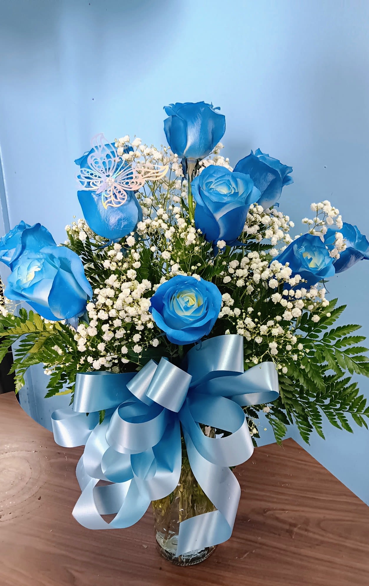AZUL - Vivid blue roses rise amid clusters of tiny white filler blooms in a clear glass vase, creating a calm, elegant bouquet perfect for a birthday bouquet, new baby celebration, get well wish, or a gentle sympathy arrangement. The cool blue and white palette feels both contemporary and soothing, the kind of gift that makes the person you're thinking of feel seen and comforted.  Stems are gathered upright so the blooms present a bold, structured silhouette while the glass vase shows the clean stems and water below. A large looping blue satin ribbon bow around the vase and a delicate pale butterfly pick add a decorative, whimsical finish that suits occasions from Mother's Day and anniversaries to housewarmings, graduations, and holidays like Easter or Christmas. This arrangement arrives ready to display; refresh the water every few days and trim stems if needed to keep the flowers looking their best.
