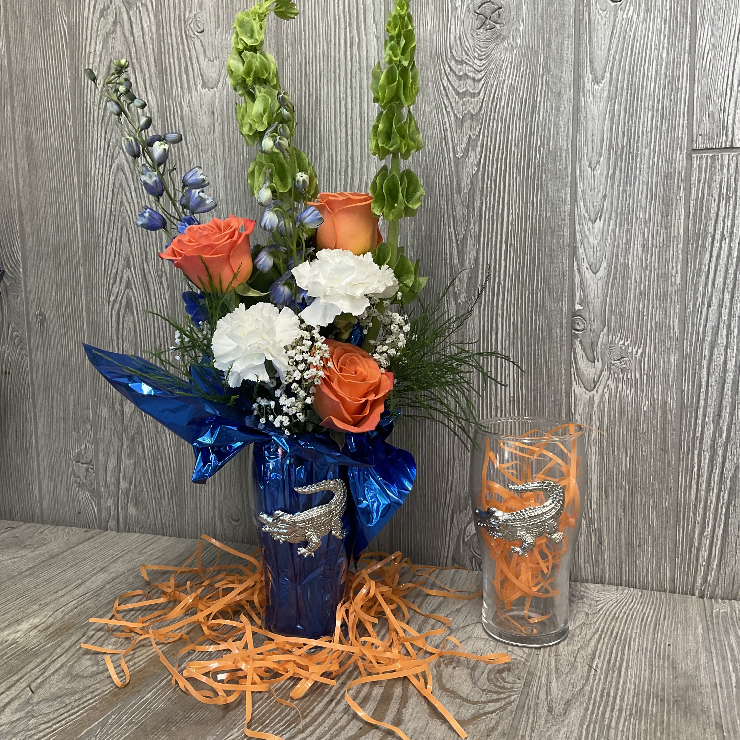 Pint Glass Gator Arrangement - Bright, playful and unmistakably spirited, this pint-glass gator arrangement is a lively choice for game day, a graduation celebration, or to send spirited congratulations. The flower arrangement brings together orange roses and white carnations with delicate white filler and tall blue-green accent stems so the person you're thinking of will feel cheered and noticed the moment it arrives. The high class pint glass by Arthur Court remains a lovely gift and reminder of good times past for years to come.  Arranged in a pint glass with shiny metallic paper, the bouquet's warm orange blooms pop against the cool blue shimmer. Perfect for fans, grads, or anyone who loves bold color, this arrangement offers a compact, eye-catching centerpiece that reads bright and celebratory in any room