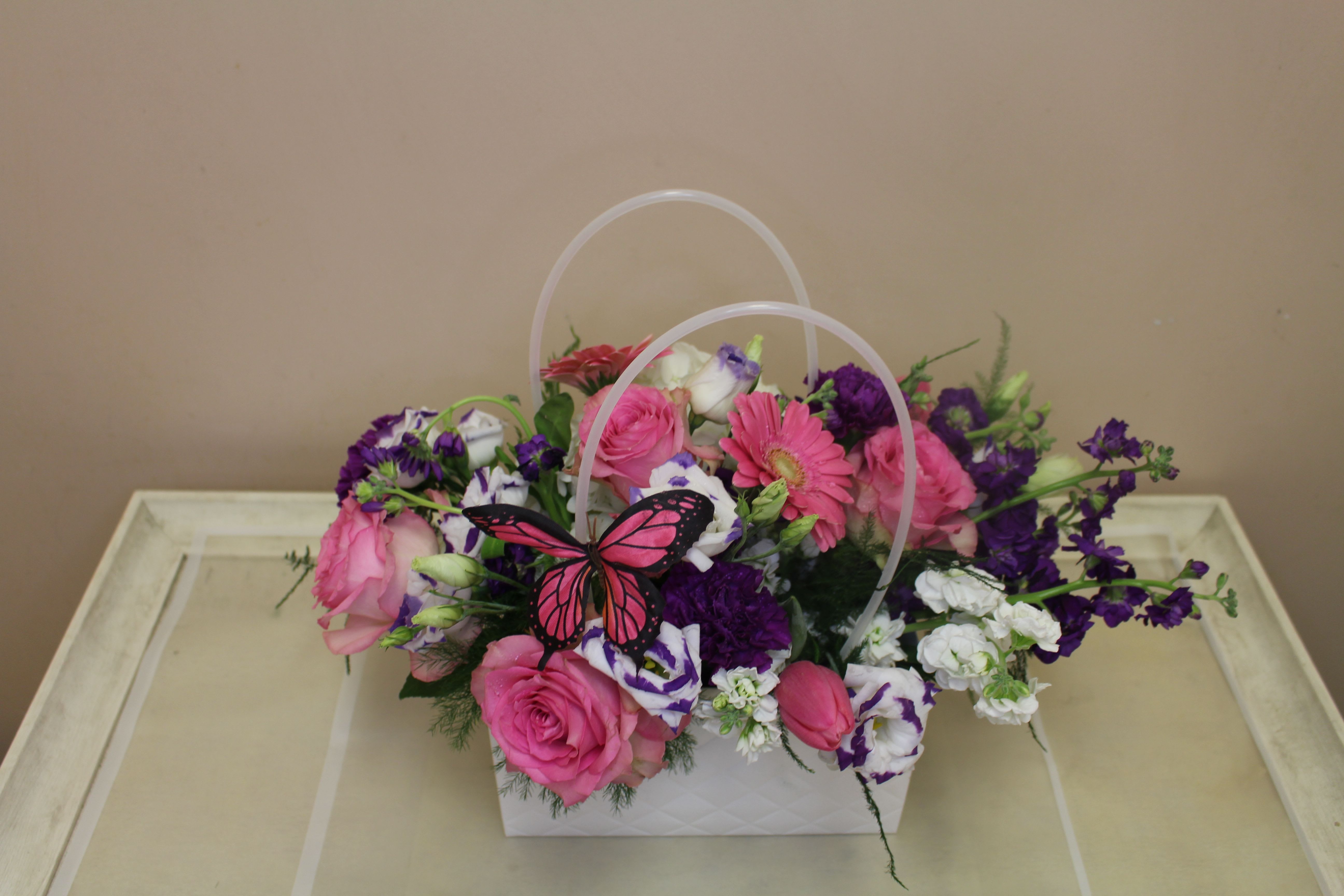 Pocketbook full of Posies Vivid - A bright decorative like pocketbook full of flowers, this floral arrangement is a cheerful gift bouquet perfect for a birthday, thank-you, or spring celebration. The mix of hot pink roses and a pink gerbera daisy gives an immediate feeling of warmth and affection, while pops of purple and white soften the palette-making the recipient feel genuinely celebrated and seen.  Arranged in a white handled decorative pocketbook, the design layers full rose heads with purple lisianthus, carnations, stock and small clustered filler blooms for a lush, rounded silhouette with a loose, abundant finish. A playful pink-and-black butterfly pick rests near the center as a decorative accent. Colors include pink, purple, white and fresh green foliage; sizing is a full. Perfect for a friend, mom, or coworker who loves bright, feminine florals, this arrangement sends a joyful, thoughtful message without being over-the-top. Order for same-day local delivery when available.