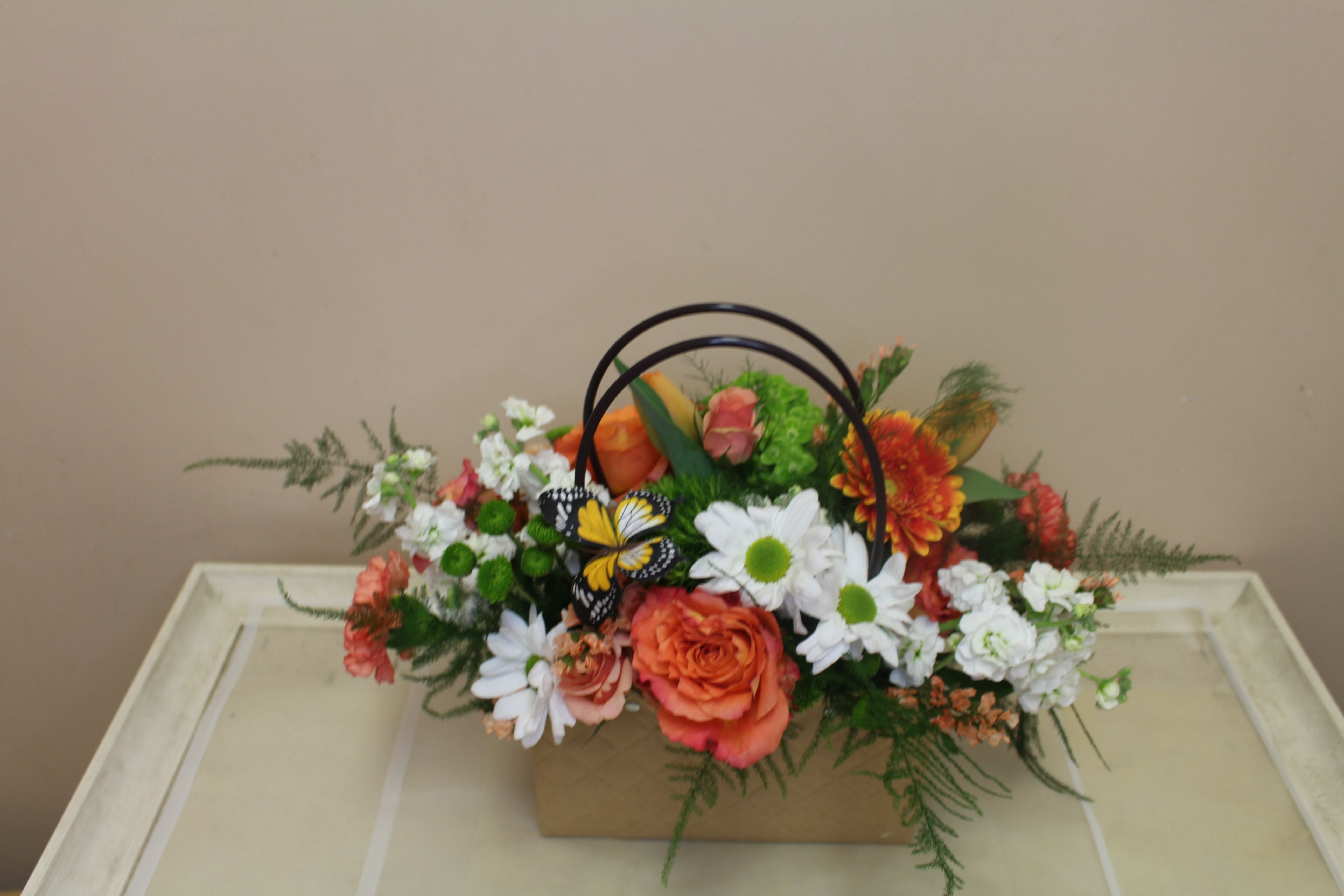 Pocketbook full of Posies bright - Bright orange roses paired with cheerful white daisies and carnations give this everyday mixed bouquet a sunny, celebratory feel - perfect as a birthday bouquet, a thank-you gift, or a "just because" surprise. Presented low and full in a rectangular decorative pocketbook-like container with a dark handle, the Pocketbook full of Posies bright reads as casual and gift-like; the little decorative butterfly adds a playful, whimsical touch that will make the person you're thinking of smile.  The arrangement mixes orange chrysanthemums and green button mums with airy filler and fern-like greenery for layered texture and a garden-style, informal composition. It's compact and horizontal, designed to sit on a table or desk without towering stems; This lively color palette of orange, white, green, and makes the recipient feel genuinely celebrated - a fresh, approachable bouquet that arrives ready to display with minimal care.
