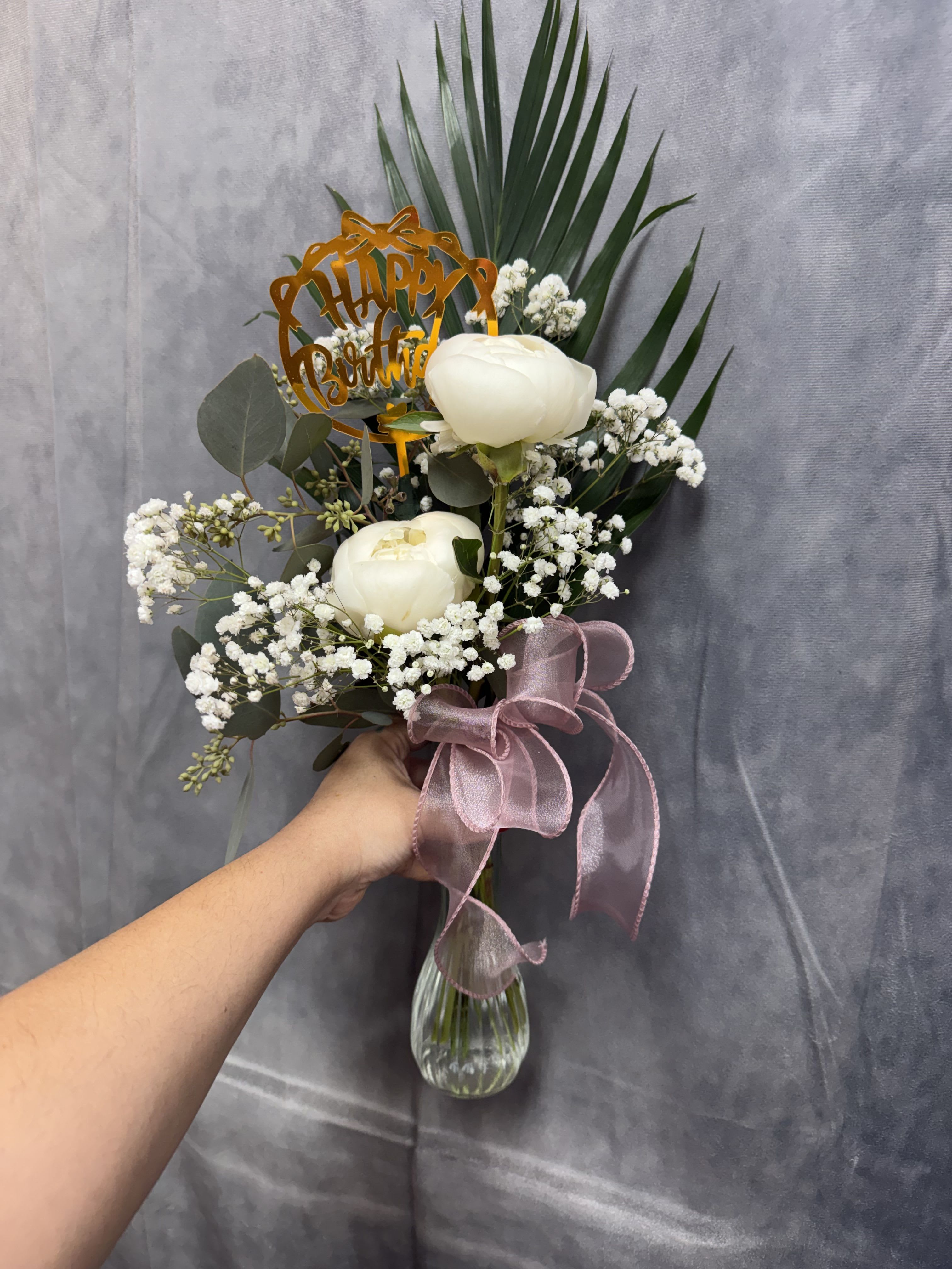 Peony Wishes Bud Vase - A soft and elegant arrangement featuring creamy peonies, delicate baby’s breath, and fresh greenery, finished with a blush ribbon and a sweet birthday accent. Simple, graceful, and full of charm—perfect for celebrating life’s special moments with a touch of timeless beauty.