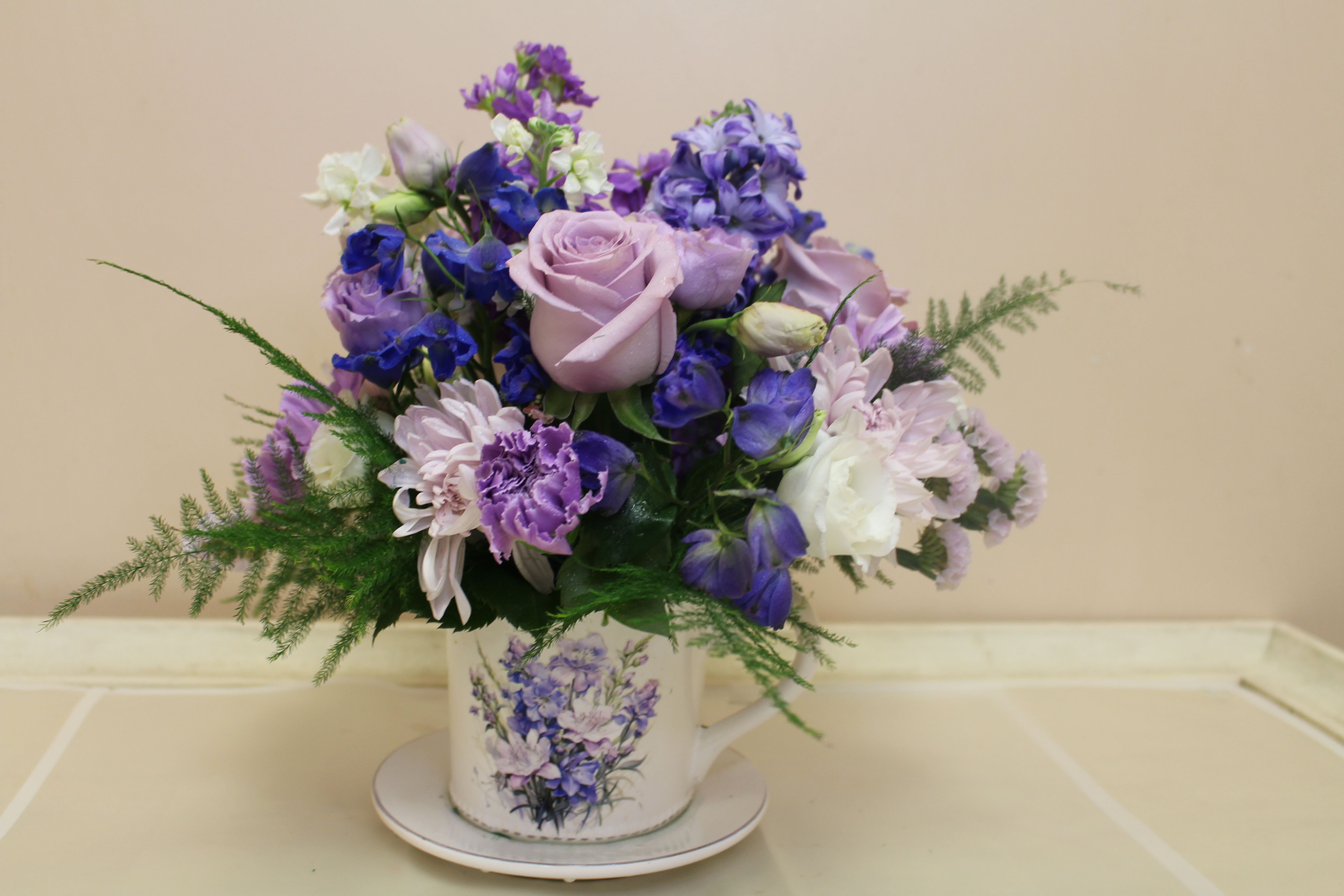 flowered coffee set - Lavender roses take center stage in this romantic teacup arrangement, a compact birthday or thank-you bouquet that reads as quietly elegant and thoughtfully feminine. Designed for occasions like birthdays, anniversaries, or a gentle "thinking of you" moment, the soft mix of purple, blue, and white blooms creates a serene, heartfelt impression that will make the person you're thinking of feel comforted and cherished.  Clusters of roses, stock, chrysanthemums, daisies, carnations, and sprays of dianthus form a rounded, layered composition with airy fern-like greenery to give a loose garden feel. The blooms are arranged low and close for a full, textured look and presented in a white ceramic teacup-style vase (with matching saucer). Its petite scale and pastel-to-saturated color palette make it a delightful gift that brightens small spaces and shows careful thought in every blossom.