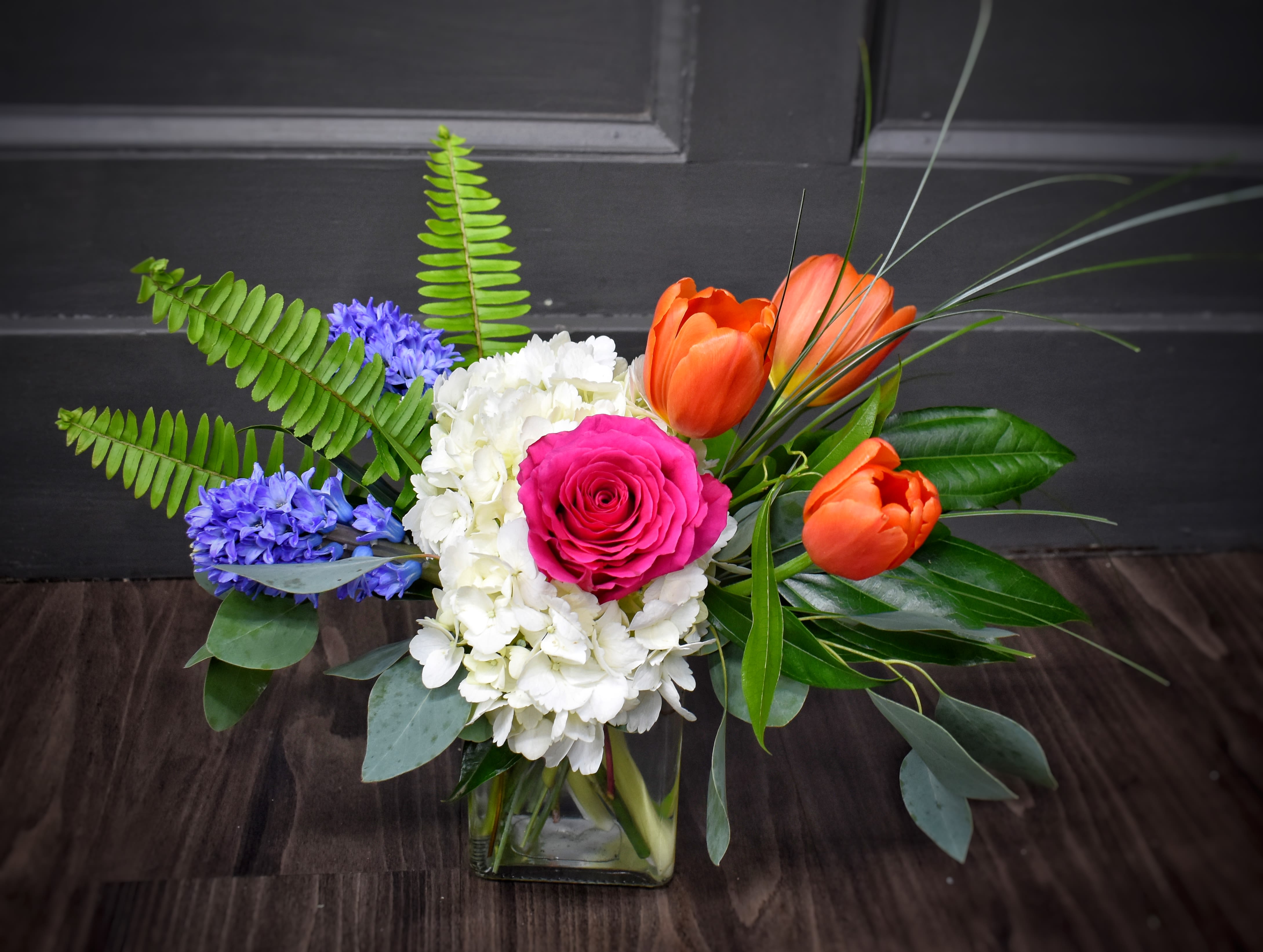 Meadow Fresh - Bright, garden-fresh color greets the recipient with a lively mix of pink, orange, yellow, purple and white-perfect as a birthday bouquet, congratulations arrangement, or an everyday celebratory gift. Meadow Fresh is a mixed-color flower arrangement designed to make someone feel truly seen: the bright pink roses read as affectionate and joyful, while the contrasting cool tones add a fresh, uplifting balance.  This full, asymmetrical design is arranged in a clear square glass vase and pairs clustered white hydrangea and daisies with orange and yellow tulips, lilies, lavender-blue hyacinth-like blooms, and mixed-color accents. Broad eucalyptus and glossy fern fronds spill outward with decorative grass for movement and texture, creating a wide, airy silhouette that highlights the soft petals against structured greenery.  Ideal for moms, friends, or coworkers who appreciate a vibrant, garden-style look, Meadow Fresh brightens a kitchen table, office desk, or entryway and sends a cheerful message for birthdays, spring celebrations, or simple thank-yous. Arrives ready to display in the included clear glass vase; trim stems and refresh the water every few days to extend bloom life.