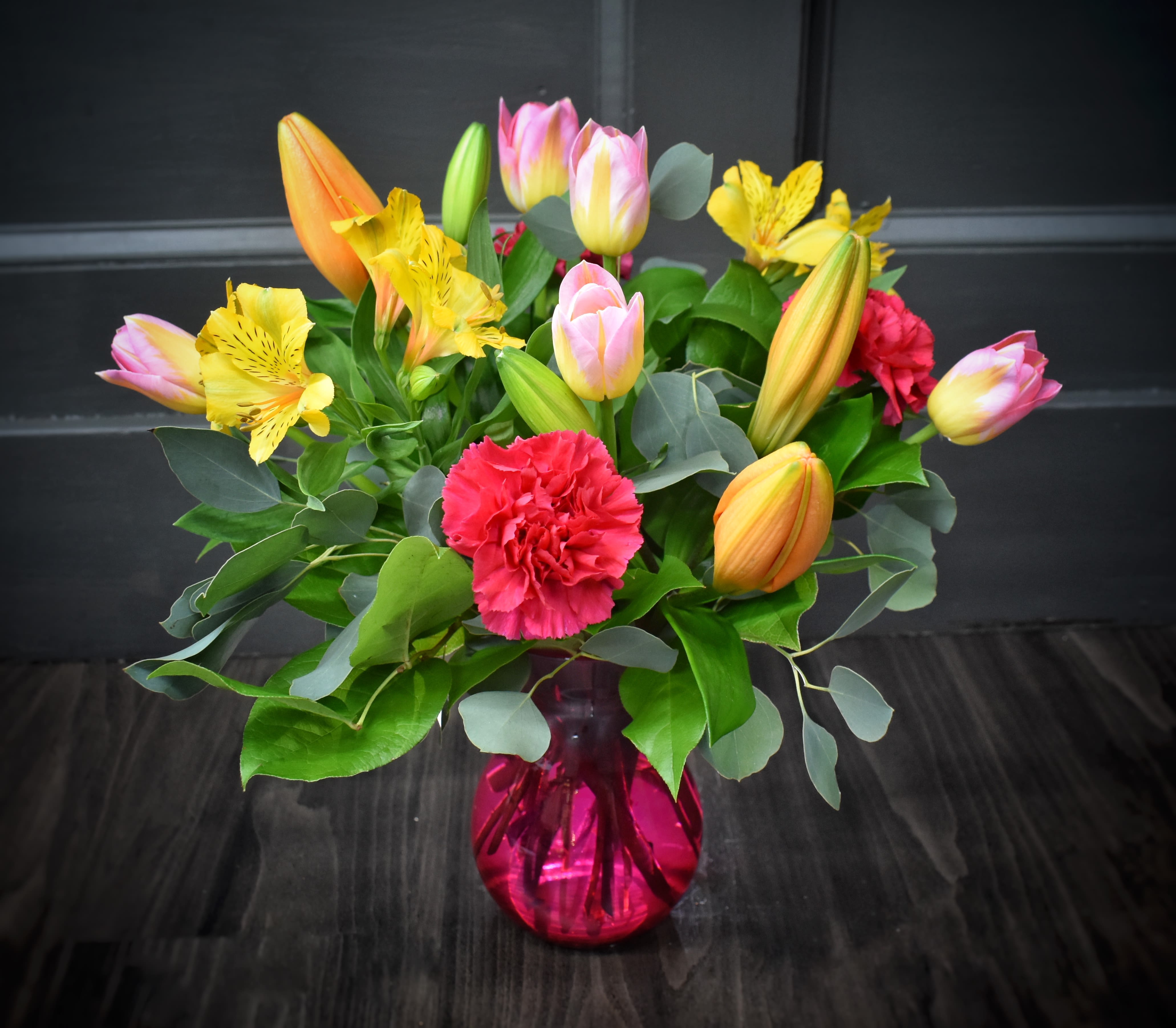 Sunny Disposition - Sunny Disposition is a cheerful mixed flower arrangement perfect as a birthday bouquet, a springtime thank-you, or an everyday way to make someone feel seen. Bright pink tulips and sunny yellow alstroemeria lead the composition, joined by orange lilies, roses, and pops of red and pink carnations for a lively, celebratory color story that reads fresh and giftable.  The flowers are arranged in a glossy pink (or, in some versions, red) glass vase for a modern, playful touch. Broad gray-green eucalyptus-like foliage frames the rounded, compact design and adds cool contrast to the warm oranges, yellows, and pinks. The overall size is a full, rounded bouquet-designed to look polished on a mantel, desk, or dining table.  Give this to a friend celebrating a birthday, send as a cheerful thank-you, or brighten someone's day during the spring season-the kind of bouquet that makes the recipient feel genuinely celebrated. Vase-ready and arranged for immediate display; light flower care (fresh water and a clean vase) will keep the blooms looking their best.