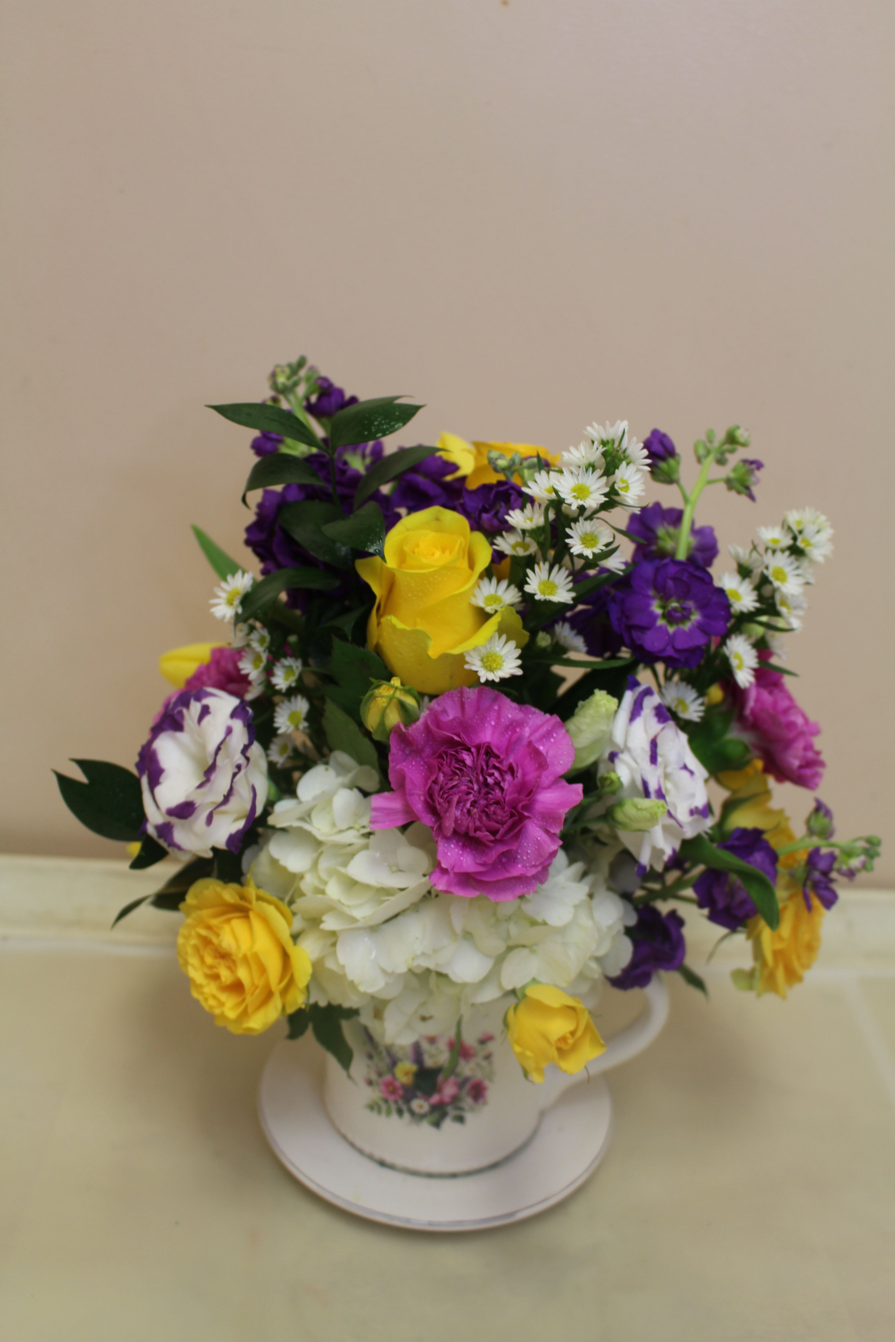 mug coffee set Bright - A bright, playful floral arrangement presented in a ceramic mug and saucer - perfect as a birthday bouquet, thank-you gift, or just-because surprise. The compact flower arrangement mixes sunny yellow roses with vivid purple and pink blooms and soft white hydrangea for a saturated, celebratory palette that still feels fresh and springlike. The teacup vase gives the gift a handcrafted, feminine charm that makes the recipient feel delightfully seen.  Cushions of small white daisy-like filler flowers and delicate lisianthus and carnations add texture and gentle contrast to the bold focal roses and magenta accents. This rounded, densely layered design sits neatly in the decorative cup (saucer included), making it an easy, display-ready centerpiece for a bedside table, desk, or kitchen windowsill. Sized for intimate gifting, it arrives ready to enjoy with simple care: keep water fresh and place away from direct heat to extend bloom life.