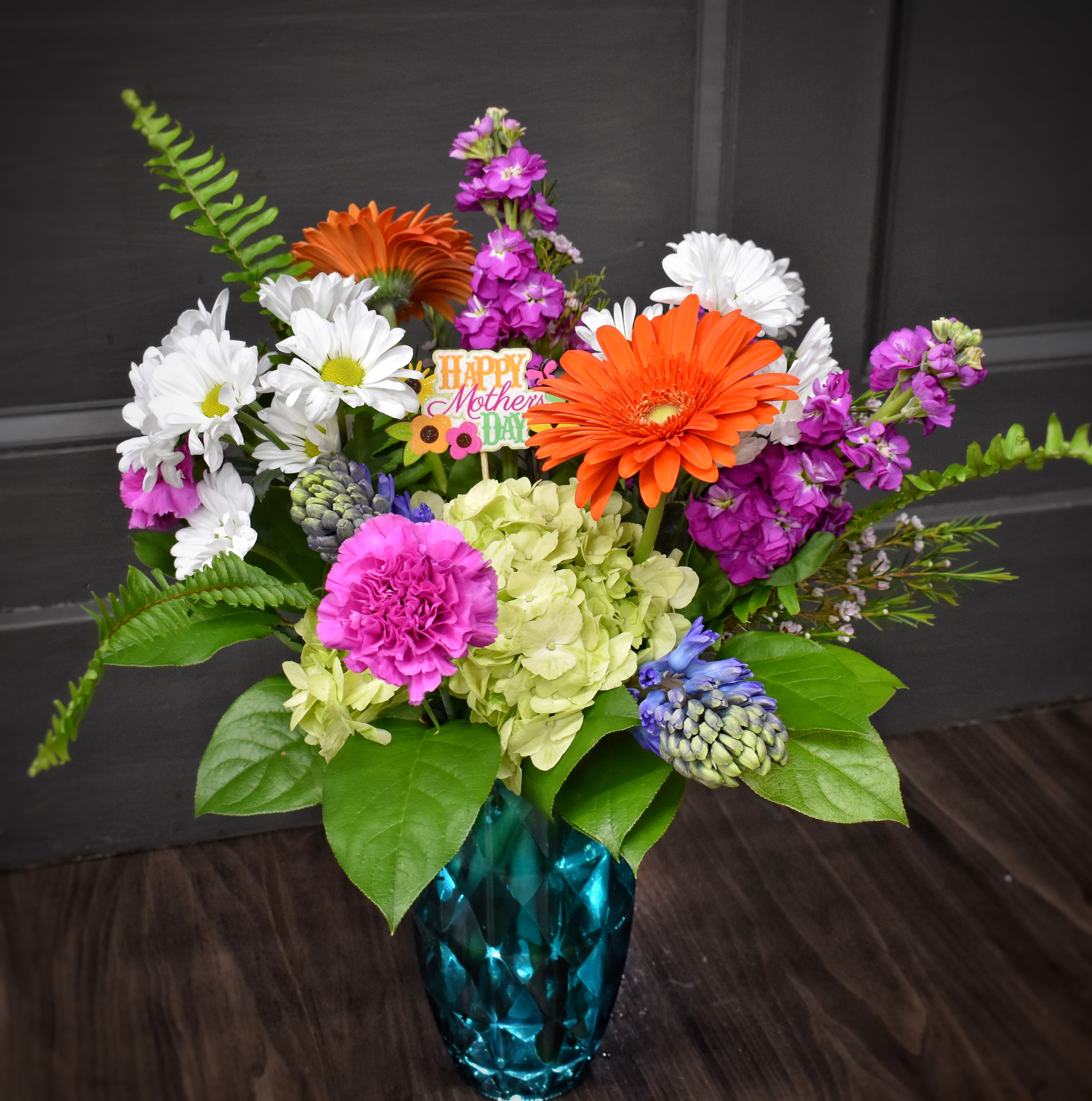 Sunshine and Smiles - A bright burst of spring-Sunshine and Smiles is a mixed flower arrangement perfect for Mother's Day or any occasion when you want her to feel celebrated. The arrangement pairs bold orange gerbera daisies and roses with soft pink carnations, white daisy blooms, pale green hydrangea, purple accent flowers and cheery tulips to create an energetic, joyful gift that reads like a hug in color.  Hand-gathered into a textured teal-blue faceted glass vase, the design balances rounded hydrangea clusters and roses with spiky, lively stems for a layered, abundant look. Broad green leaves frame the blooms and a small Mother's Day pick is tucked into the center to announce the occasion. Sized to feel generous on a table or counter, this arrangement arrives ready to display; change the water every couple days and trim stems slightly for longest freshness.