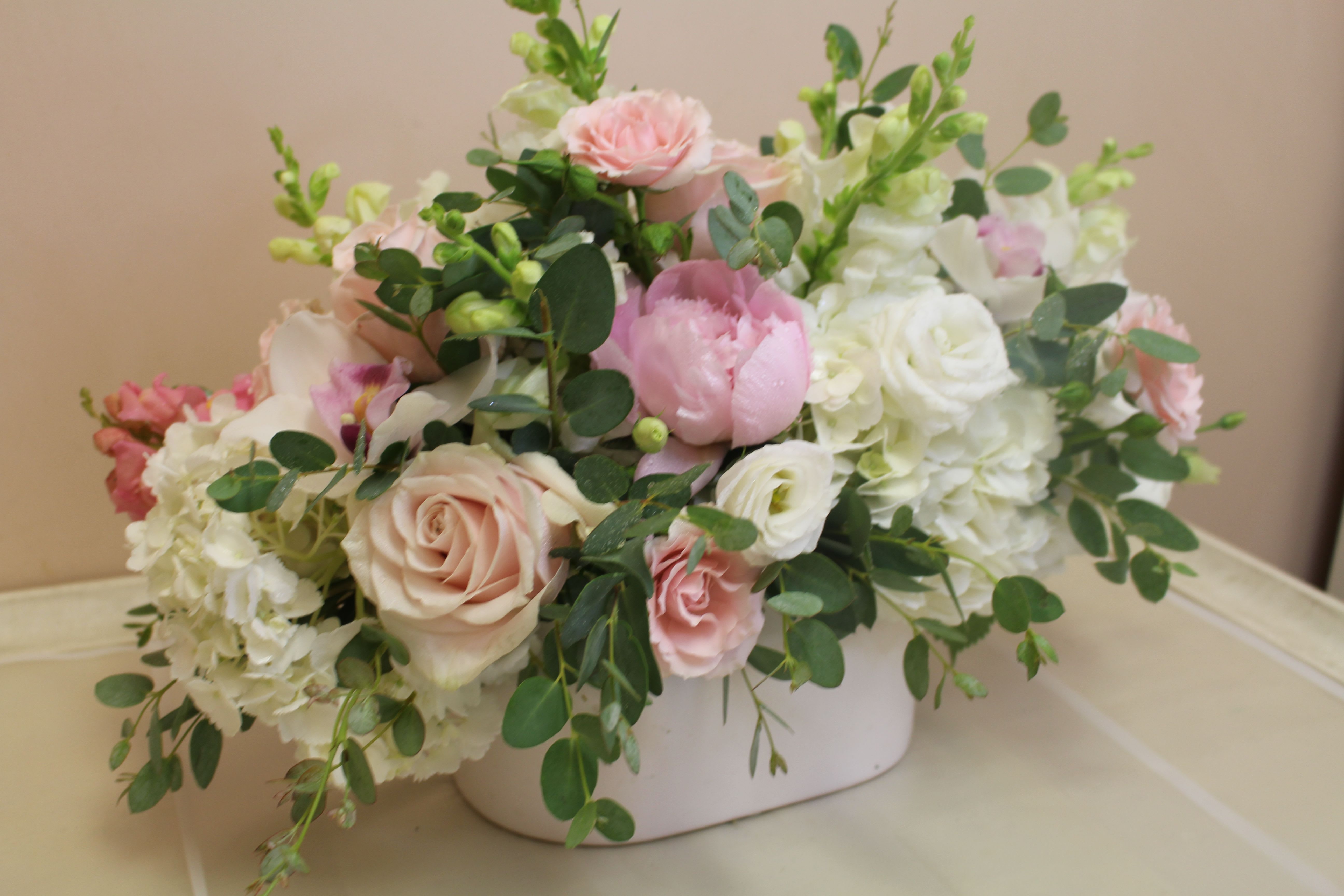 Lush Romance - Soft blush roses and ruffled peonies nestle among creamy white hydrangeas and lisianthus in a low, wide centerpiece made for weddings, anniversaries, or any moment that calls for a gentle, romantic touch. This garden-style flower arrangement reads as a tabletop centerpiece or mantel display-the soft pinks and whites, lifted by airy greenery, create a calming, elegant presence that will make the person you're celebrating feel truly cherished.  The arrangement is crafted in a simple white ceramic container that keeps the focus on the blooms and complements formal settings. Rounded clusters of roses, peony petals, hydrangea heads, tulip and spray stock accents, plus delicate filler flowers and trailing greenery, give the piece lush volume while remaining compact and horizontal in shape. Ideal for a reception table, bedside tribute, or intimate anniversary dinner, it arrives ready to display-no vase needed-and will hold its garden-fresh look when kept in a cool spot with fresh water.