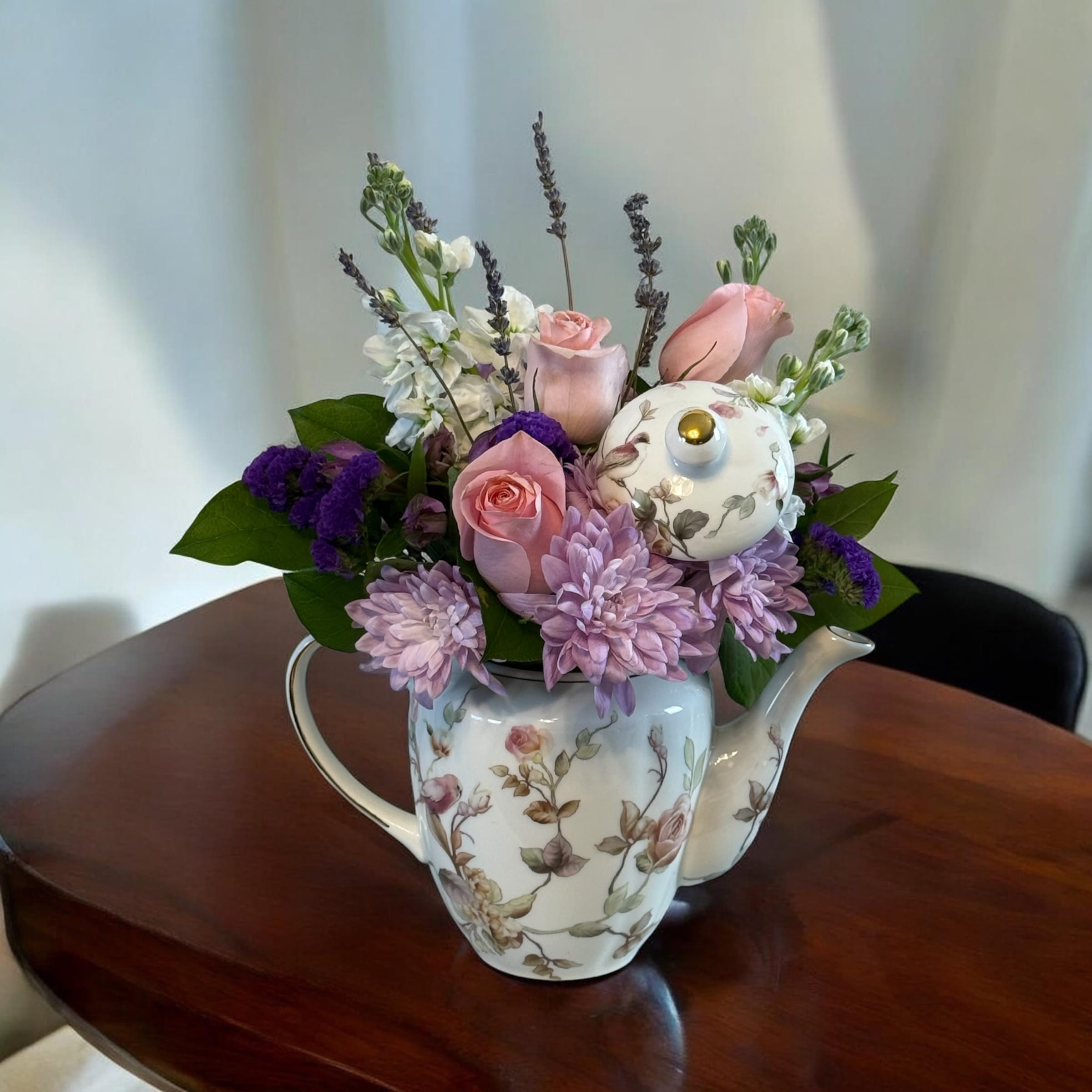 Tea time with Mom by Petal Perfection Flower Company - A vintage-inspired mix of soft pink roses and lavender chrysanthemums arranged in a white ceramic teapot makes this flower arrangement a perfect birthday bouquet, Mother's Day gift, or "thinking of you" surprise for Mom. The teapot container and decorative floral lid give the arrangement a whimsical tea-party feel that will make the person you're celebrating feel cherished and remembered.  Creamy white stock and delicate purple filler flowers add height, fragrance, and texture to the layered pastel palette of pink, purple, white, and green, while a few deeper violet accents create gentle contrast. The compact tabletop design works well on a kitchen table or mantle-an everyday centerpiece that reads like springtime charm. Care: keep stems in fresh water, trim ends every few days, and place away from direct heat for best longevity.