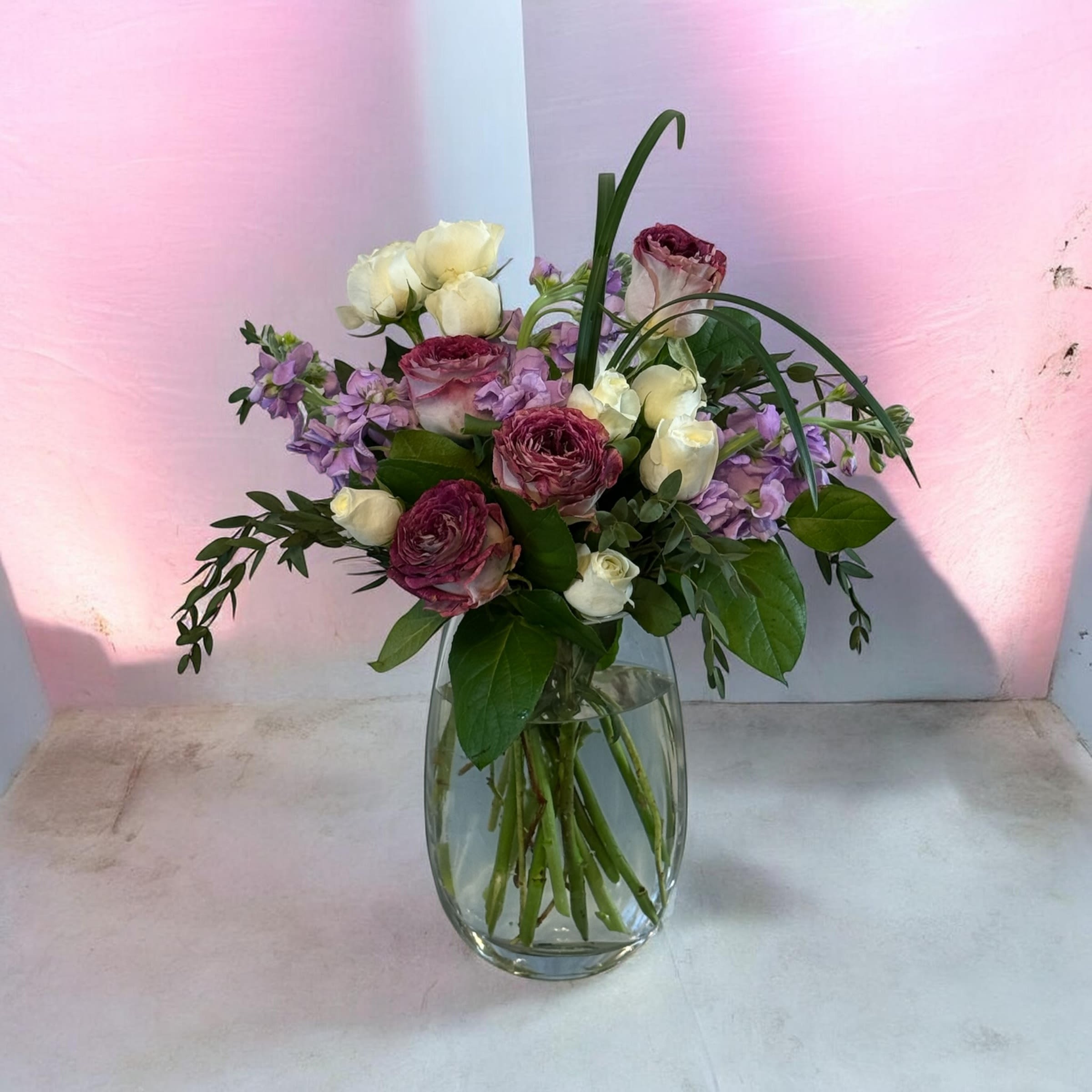 The Elegance of Motherhood Vase by Petal Perfection Flower Company  - Cream and Lavender roses take center stage in this vase arrangement, creating a soft yet slightly dramatic statement perfect for a Mother's Day, birthday bouquet, anniversary roses, or a thoughtful "thinking of you" gift. The combination of romantic cream petals and deeper wine-colored blooms, accented with lavender filler and broad green foliage, gives the person you're sending it to a feeling of being cherished and noticed.  Arranged in a clear "flat" glass vase with stems visible through the water, the design reads fresh and natural-an elegant everyday arrangement that still feels special. Colors include pink-leaning cream, rich lavender, soft purple accents, and lush green leaves. The mix of roses and clustered lavender-like filler provides contrast in texture and a gentle scent that enhances the mood without overwhelming.  This piece suits parents, partners, or friends celebrating milestones or receiving comfort-an intimate way to say you're thinking of them. Vase and arrangement are delivered ready to display; trim stems and refresh water every few days to extend bloom life.