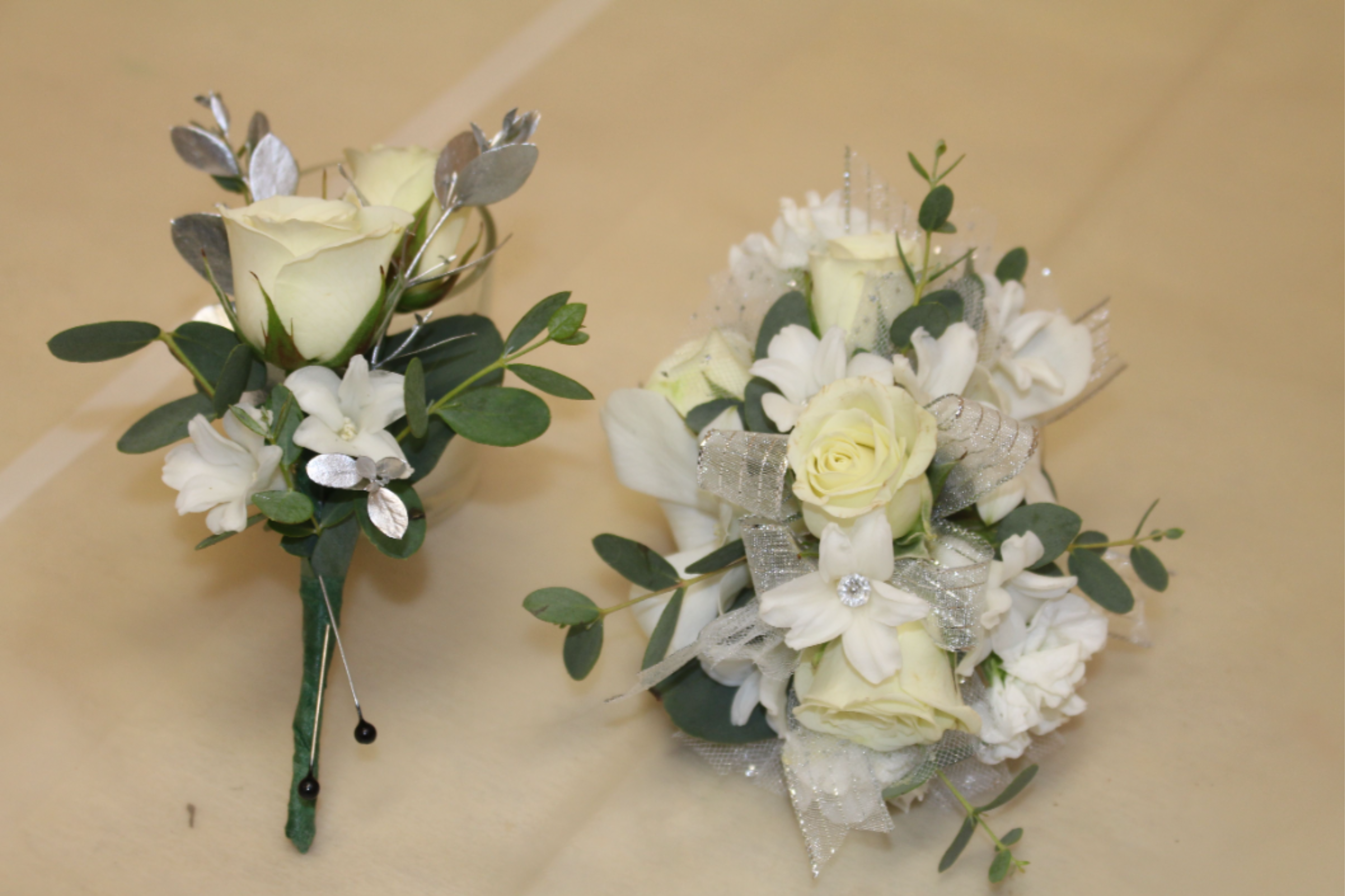 All white corsage and boutonniere bundle  - A pair of all-white corsage and boutonniere pieces that read as timeless and quietly luminous - perfect as a prom or any formal ceremony. These small floral accents combine ivory and white roses with tiny white hyacinth and rounded dark-green foliage, so the person you're honoring will look polished and cherished without anything flashy.  Each piece is finished with white and silver ribbon, decorative tulle subtle metallic leaf accents for a touch of sparkle, plus boutonniere pins for secure wear. The compact, pinned stems are sized for lapels or wristwear and keep the silhouette neat and comfortable.  Wrist corsage is on a rhinestone bracelet  