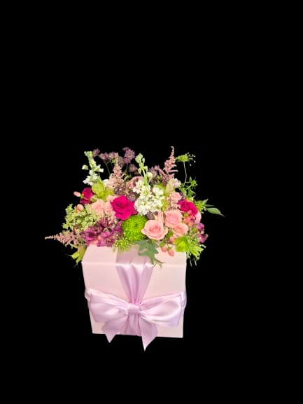 Mother's Light - A dense cluster of blush and magenta roses with white stock and green daisies makes this gift-box arrangement feel like a secret garden in bloom - perfect as a birthday bouquet, anniversary flowers, or a Mother's Day surprise. The compact, romantic presentation rises with airy snapdragons and branching stems so the recipient sees layers of texture and color the moment the box is opened.  Carefully arranged in a pale pink gift box tied with a wide satin ribbon, the mix includes roses, stock, dianthus and other seasonal filler blooms in pink, purple, white and bright green accents that keep the palette fresh and lively. The box-mounted design creates a polished, celebratory look that travels well and needs minimal fuss - no vase required, simply place on a table and enjoy the fragrant, garden-style display.