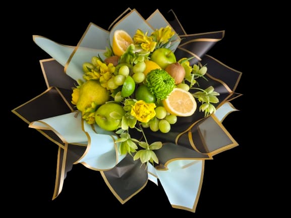 Limoncello - Bright, playful and unmistakably gift-ready, this Limoncello flower arrangement pairs cheerful yellow blooms with a bold fruit-forward twist - perfect as a celebratory bouquet or thank-you gift. The compact, rounded composition reads more like a fruit-and-flower bouquet than a traditional floral arrangement, so the person you're thinking of will feel delighted by its fresh, upbeat surprise.  Green apples, grapes, pears, citrus halves and kiwi-like fruit are tucked among small yellow daisy-like blossoms for a lively green-and-yellow palette accented with touches of white and deep blue. The stems and fruit are wrapped in layered pale blue and black paper with gold edging for a polished, modern presentation; decorative wrapping paper gives the whole piece a dramatic, gift-like finish that travels well.  Choose this arrangement to mark birthdays, congratulations, or a spontaneous "thinking of you" moment when you want someone to feel brightened and seen. The compact size makes it easy to display on a table or countertop; to keep fruit and small blossoms looking their best, place the bouquet in a cool spot away from direct sun and enjoy within a few days for peak freshness.