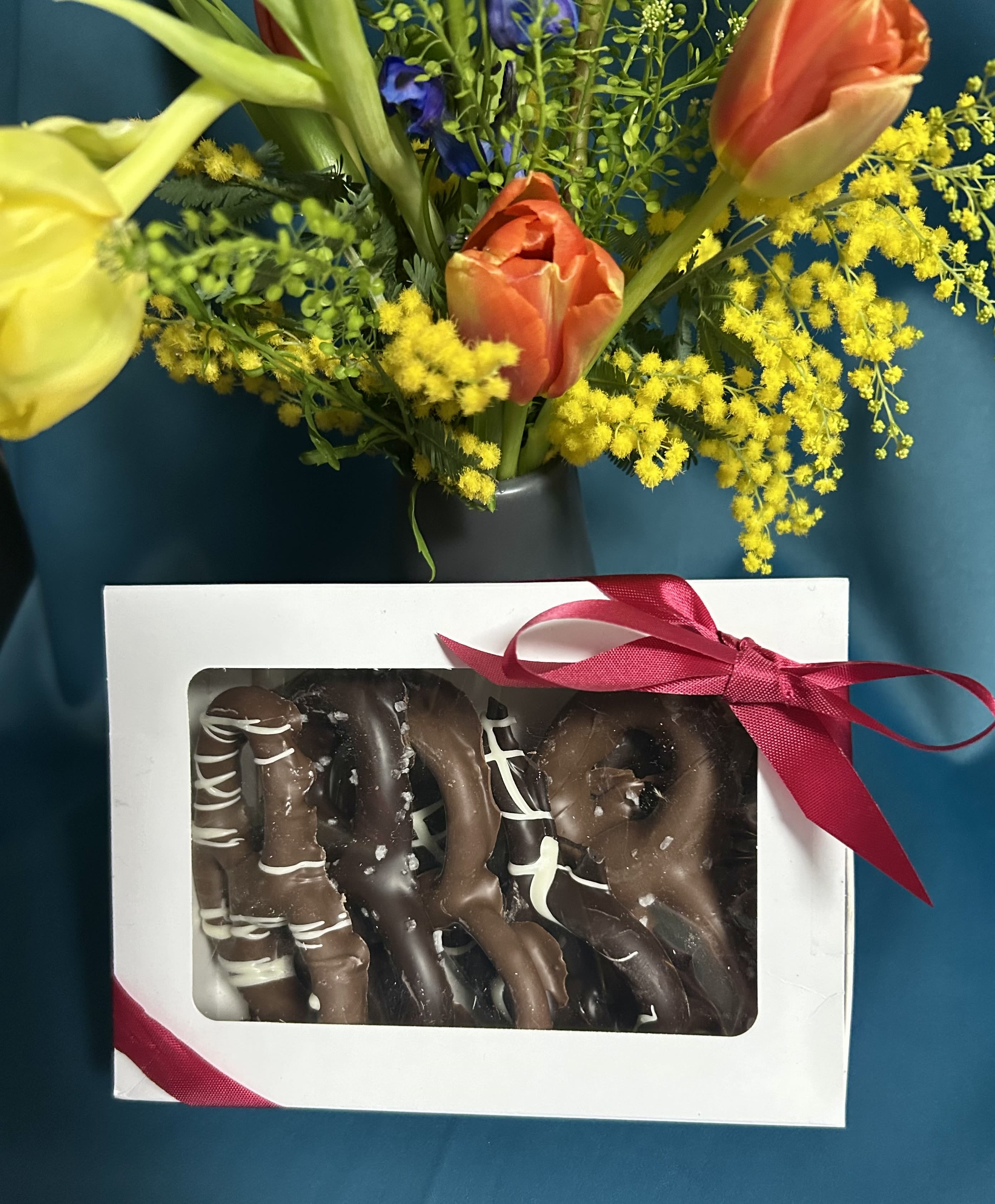 Gift set with yellow and orange tulips in a vase behind a box of chocolate-covered pretzels tied with a red ribbon.