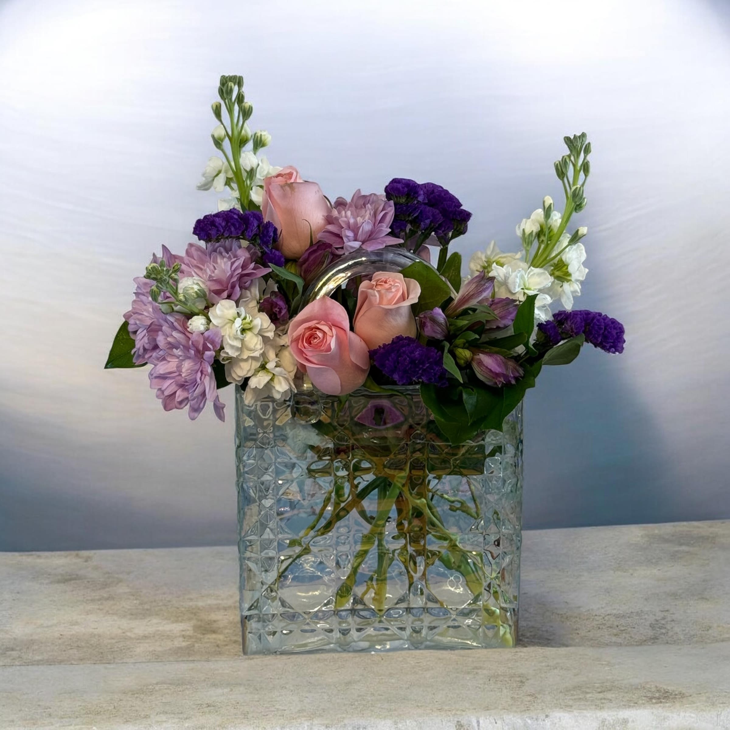 Petal Perfection's "Let me grab my purse!" - Soft pink roses and lavender chrysanthemums lead this mixed-color flower arrangement, a bouquet perfectly suited as a Mother's Day,  birthday bouquet or everyday gift to make someone feel cherished. The combination of rounded roses and carnations with upright white stock and purple accents creates a romantic, feminine look that reads fresh rather than formal-the kind of arrangement that says "you're on my mind" with a playful, elegant touch.   Clear textured glass handbag-shaped vase that adds a decorative, gift-like flair. Flowers include roses, carnations, stock, chrysanthemums, alstroemeria and complementary filler blooms in pinks, purples and white; the airy mix of shapes and heights keeps the arrangement balanced and lively. Sized for tabletop display, it's an easy-to-place surprise that will make the recipient feel genuinely celebrated-no special care beyond fresh water and a cool spot to extend blooms.